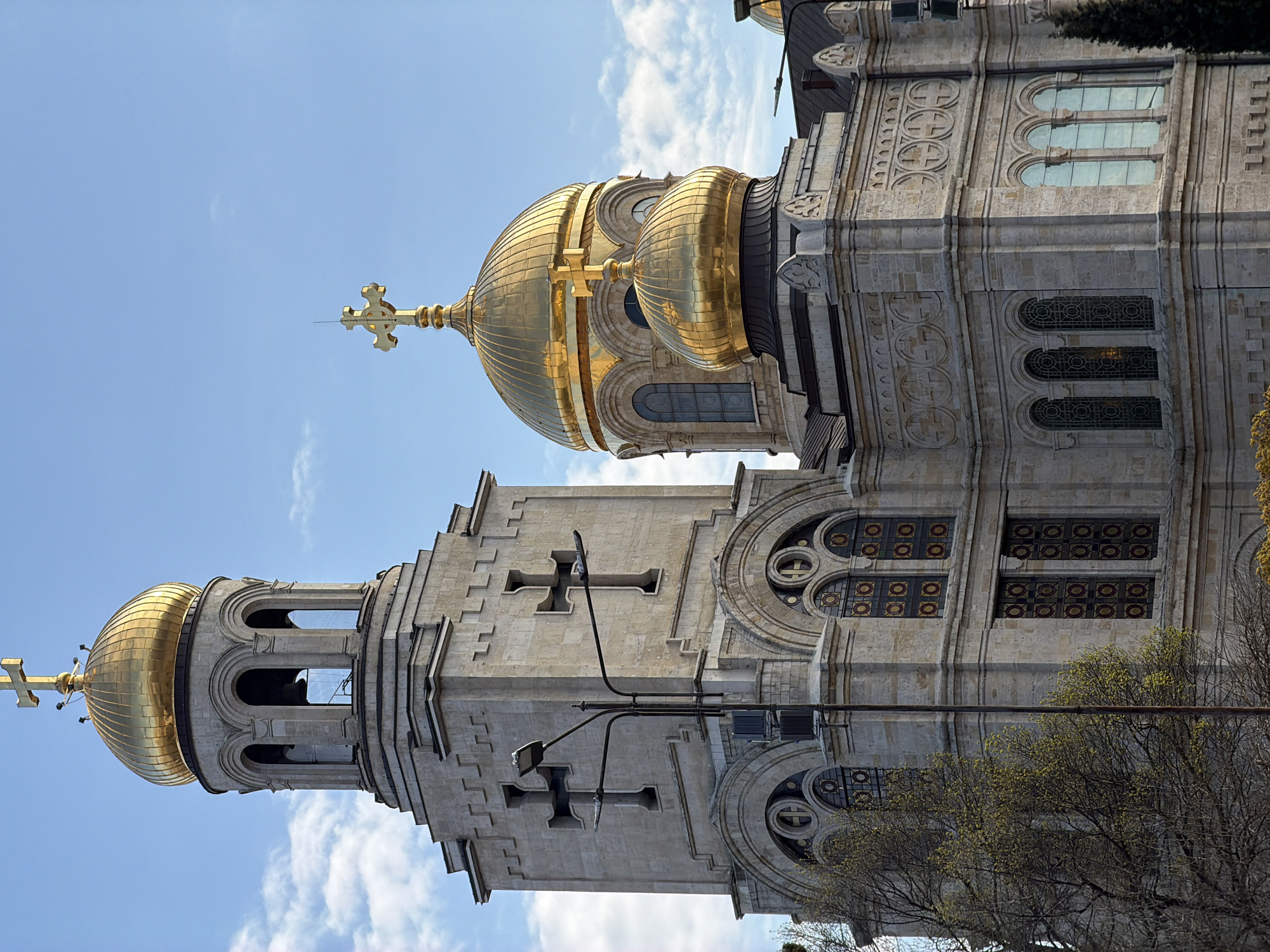 The image depicts the Dormition of the Mother of God Cathedral in Varna, Bulgaria, showcasing its architectural details and golden domes against a blue sky with clouds. The church features intricate stonework, arched windows with decorative patterns, and prominent crosses. A lamppost is visible on the side of one of the towers. Part of a tree is visible near the cathedral’s structure.