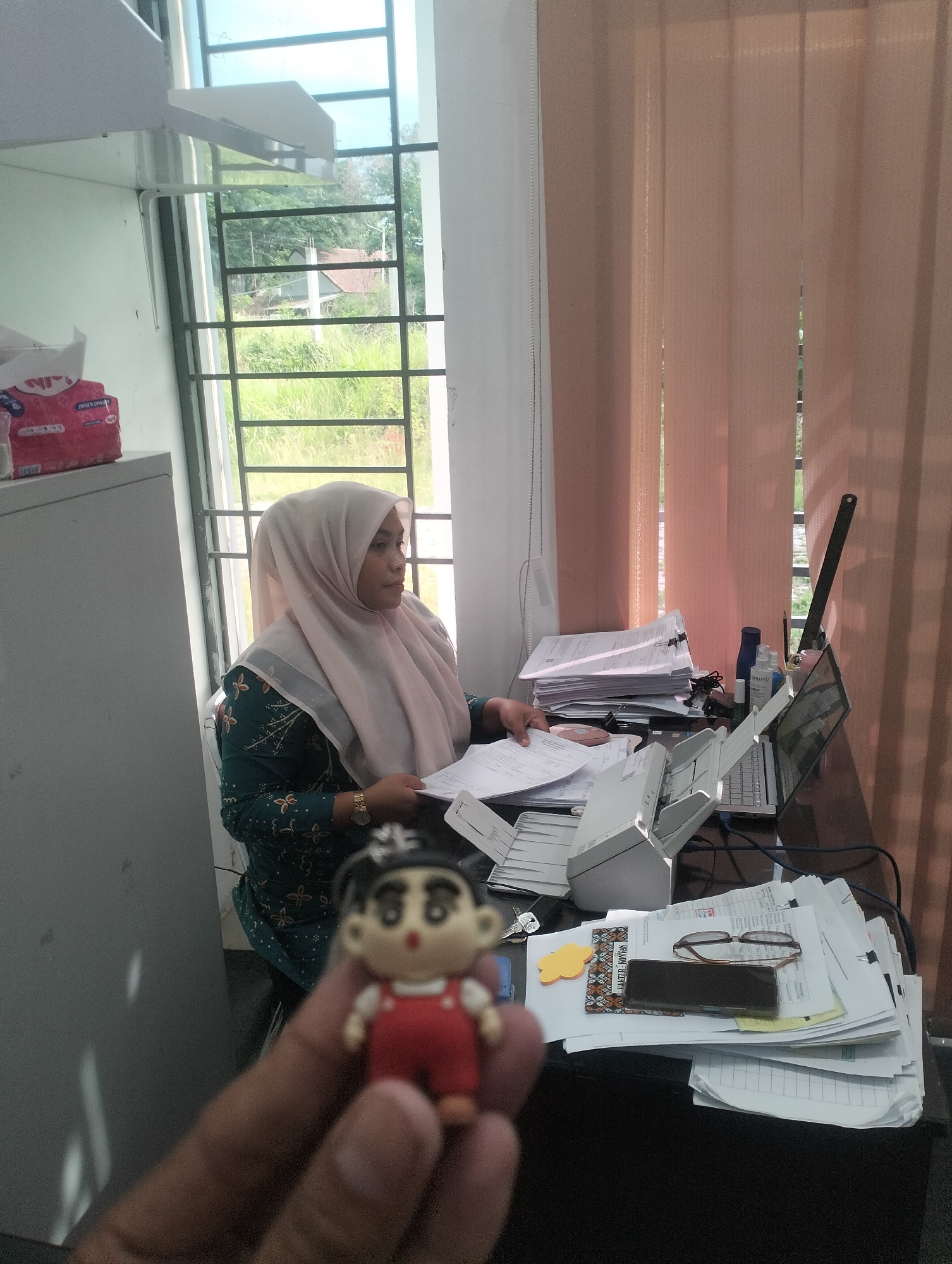 The image depicts an indoor scene, likely an office, in Lhokseumawe, Indonesia. A woman wearing a light pink hijab and a dark green patterned blouse is seated at a desk, engrossed in paperwork. Her attention is directed towards the documents in front of her. On the desk, there are stacks of papers, a scanner, a laptop, a bottle of hand sanitizer, and a pair of eyeglasses. The room features a large window with metal grates, offering a view of lush greenery and a distant building. Vertical blinds in a peach hue hang on the right side of the window. In the foreground, a blurred image of a small figurine, resembling a cartoon character, is being held up by a hand. The overall impression is one of a working environment, possibly a government office or a business, with the natural surroundings of Lhokseumawe visible through the window.