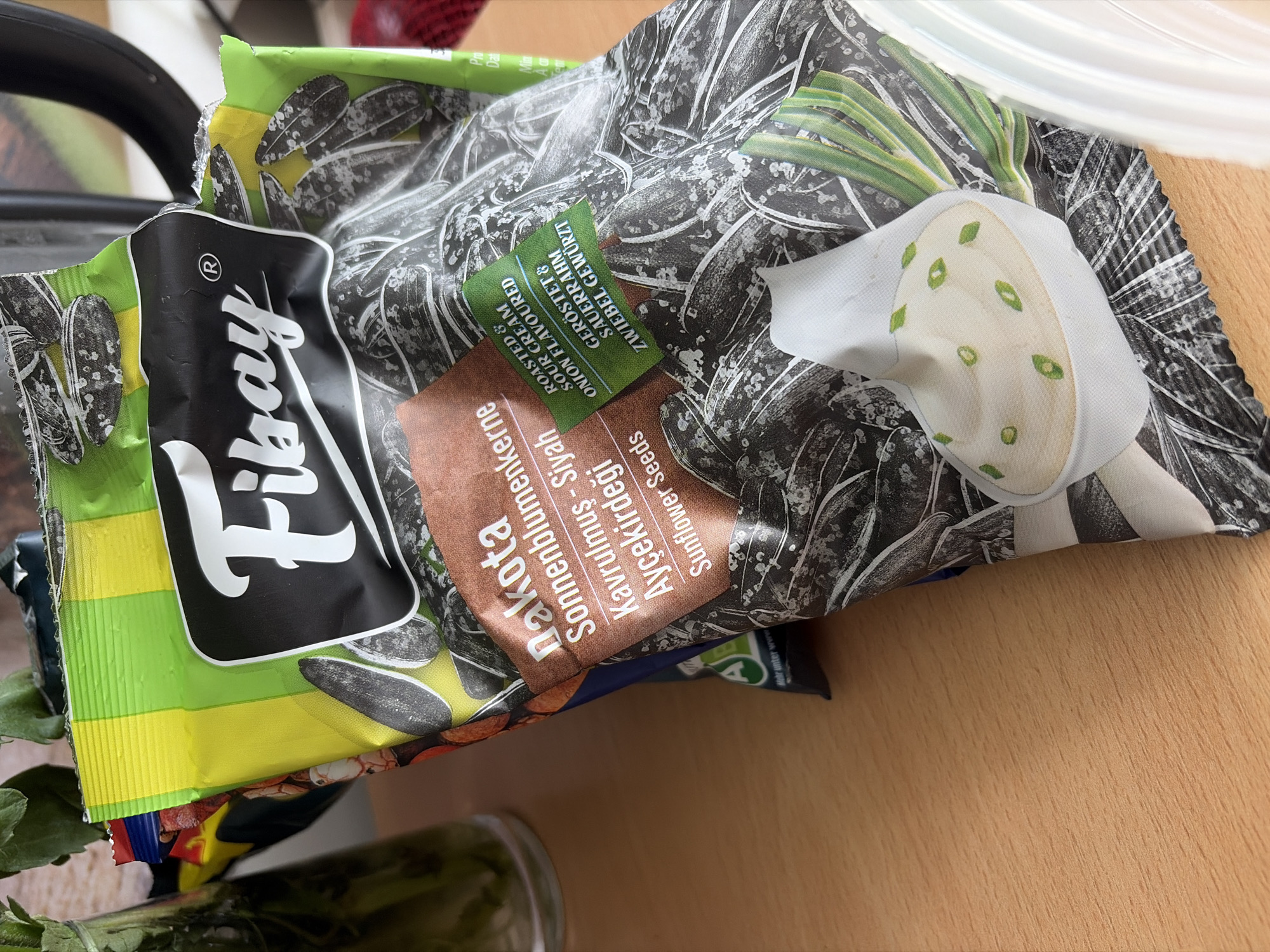 The image shows a package of "Fikray" brand sunflower seeds with a "Dakota" label. The package is predominantly green and black, with the "Fikray" logo in white text within a black oval. The "Dakota" label is a brown rectangular patch with white text. Below this, a green label indicates "Roasted & Sour Cream Onion Flavoured" and "GERÖSTET & SAUERRAHM ZWIEBEL GEWÜRZT". The packaging depicts images of sunflower seeds and a dollop of sour cream and chives. The package is resting on a wooden surface. There is a glass vase with green foliage in the upper left corner and a portion of a black appliance in the upper right corner. The overall scene suggests a domestic or retail environment, possibly a kitchen counter or a shop shelf in Duisburg, Germany. No people are visible in the image.