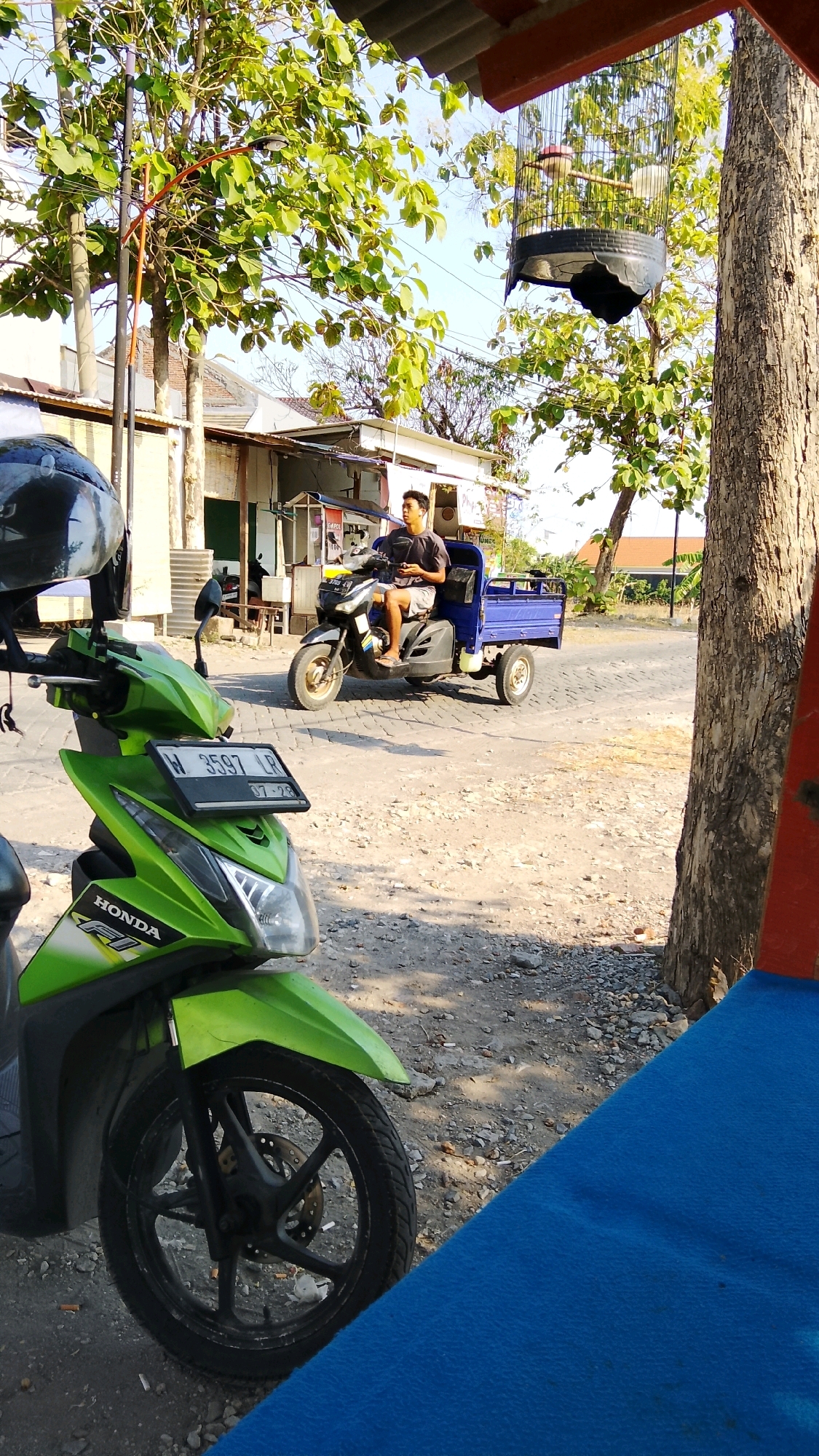 The image shows a sunlit outdoor scene, likely in Dahanrejo, Indonesia. In the foreground, a green Honda scooter is parked, its front wheel and fender clearly visible. To the right, a portion of a large tree trunk and a red structure with a blue fabric are present. In the mid-ground, a young man is riding a blue three-wheeled motorized vehicle, resembling a mini-truck, down a paved road. He is wearing a dark t-shirt and light-colored shorts. Behind him, there are small buildings with corrugated roofs and trees. In the upper right of the image, a birdcage hangs from a tree branch, containing a bird. The sky is clear and blue. The environment appears to be a roadside setting with some vegetation and urban elements.