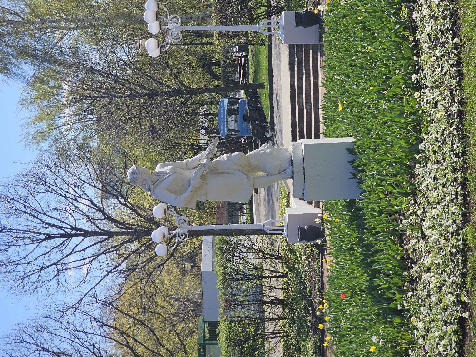 The image depicts an outdoor scene in what appears to be a park in Moscow, Russia. A white sculpture of a man in formal attire stands on a pedestal. The man is holding what seems to be a scroll or document. The sculpture is located near a flower bed filled with greenery and yellow and white flowers. Behind the sculpture, there are lampposts with multiple globe-shaped lights. In the background, there are trees, benches, a bus, and some stairs. The overall environment suggests a well-maintained park or garden area.