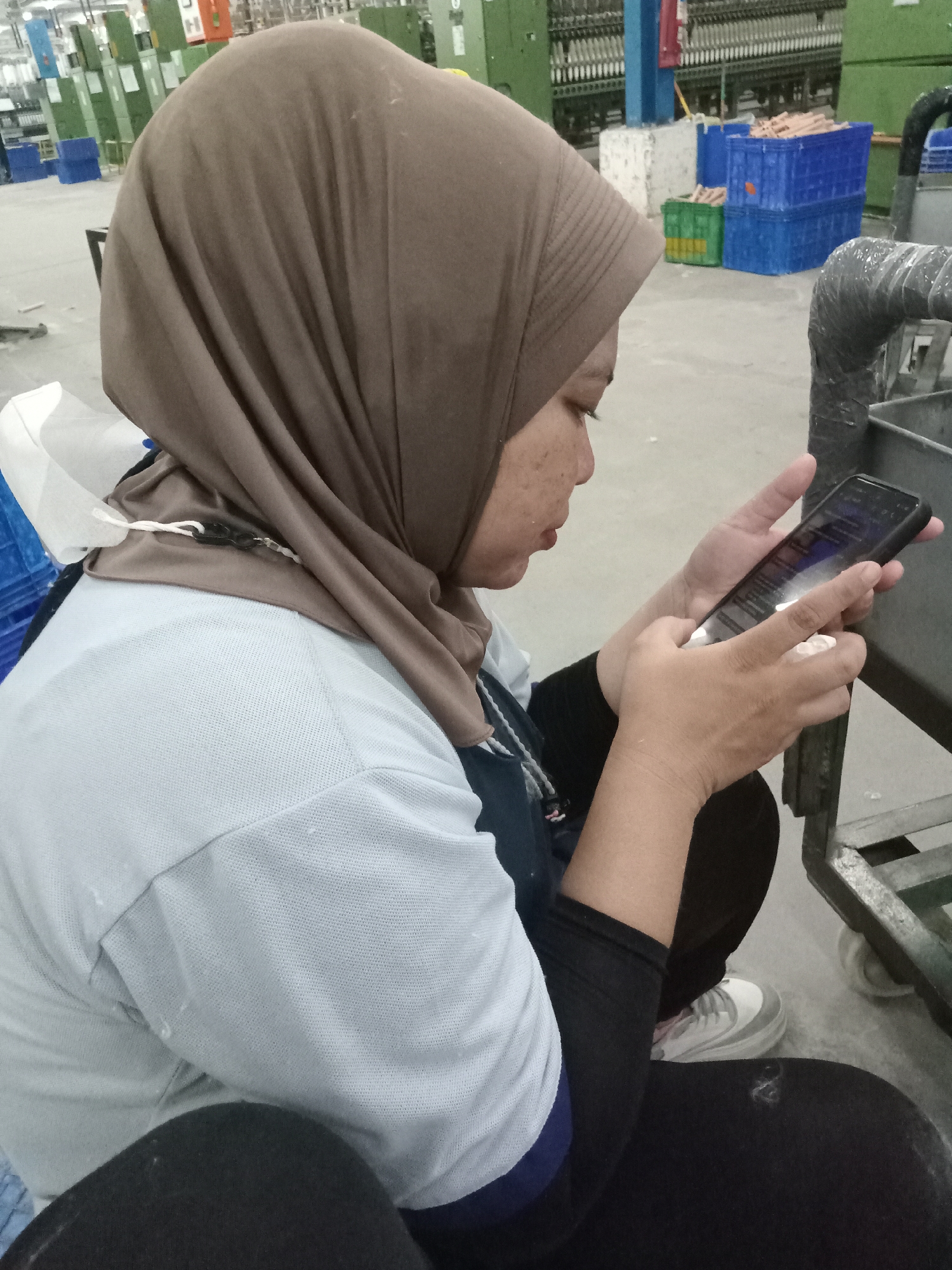 A woman wearing a light brown hijab and a light blue t-shirt with a dark blue collar is seated on the floor, looking at a smartphone. She is also wearing black leggings and white sneakers with gray accents. A white surgical mask is partially visible near her neck.
The background appears to be an industrial setting, potentially a factory, with green and blue machinery and storage bins. The location is Sukomoro, Indonesia.