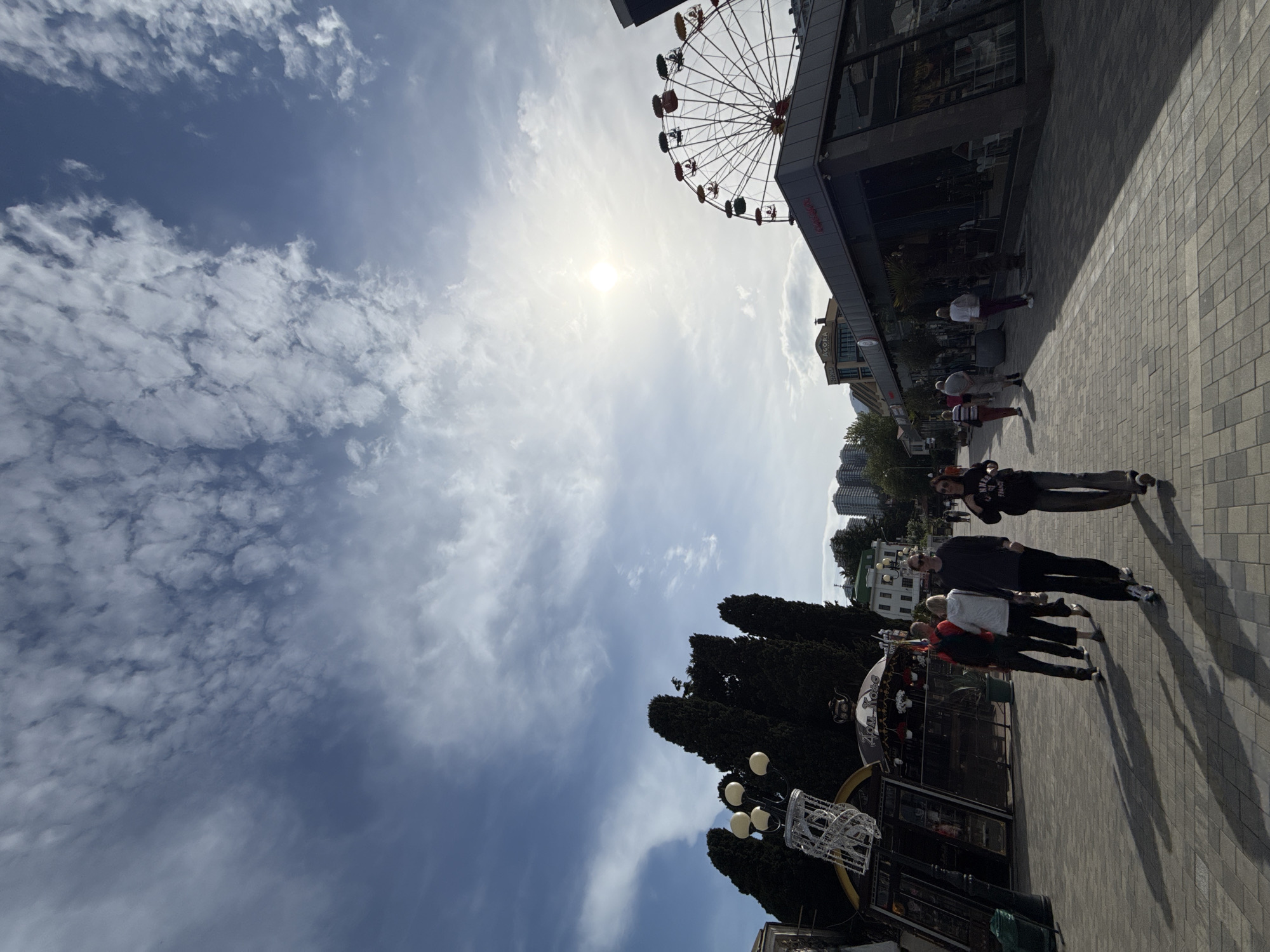 The image depicts an outdoor scene with a Ferris wheel partially visible in the upper right corner against a bright, cloudy sky with the sun positioned near the top center. The foreground shows a paved walkway where several people are walking. There are trees and building structures visible along the side of the walkway. The location is in the city of Unknown, Unknown.
