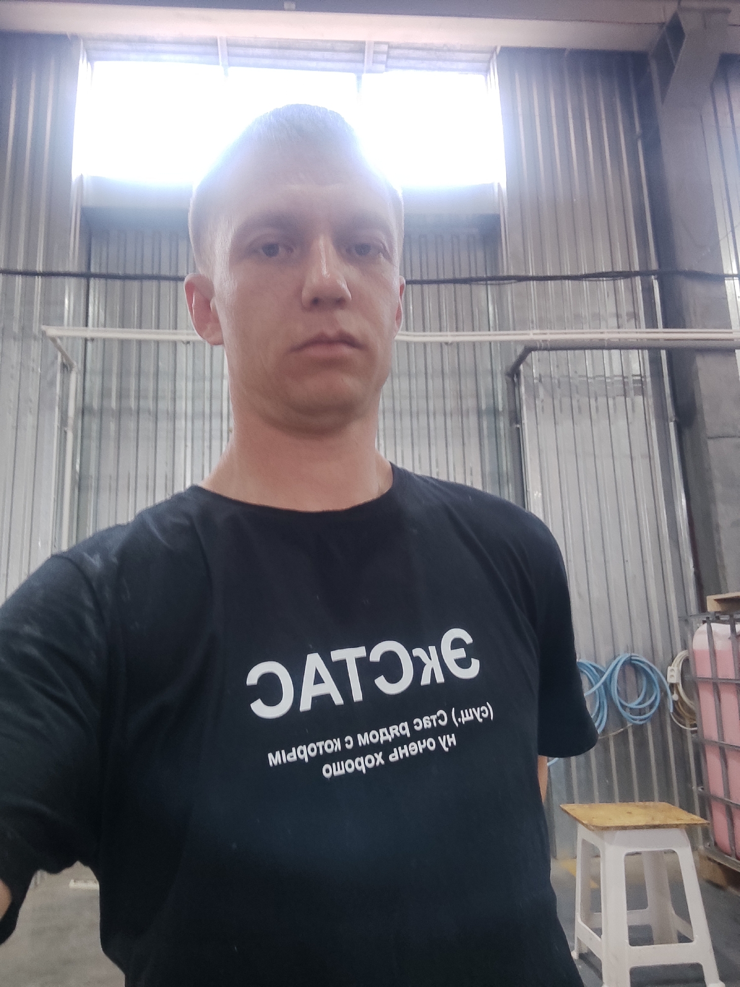A man stands in what appears to be a factory or industrial setting, looking directly at the camera. He has short hair and a serious expression. He is wearing a black t-shirt with white text printed on it. The text on his shirt is in Russian and appears to be in a stylized font, with "ЭКСП" clearly visible in large letters at the top. Below this, there is smaller text, also in Russian.
The background shows a large, open space with corrugated metal walls and a bright, possibly natural, light source at the top, suggesting a window or skylight. Metal structures and pipes are visible, along with some industrial equipment such as coiled blue hoses and a pink container. A small white stool with a wooden top is positioned to the right of the man. The overall impression is of an indoor industrial environment. There are no clear indicators of the time of day or weather, but the lighting suggests it is daytime. The man's expression is neutral, conveying no strong emotions.