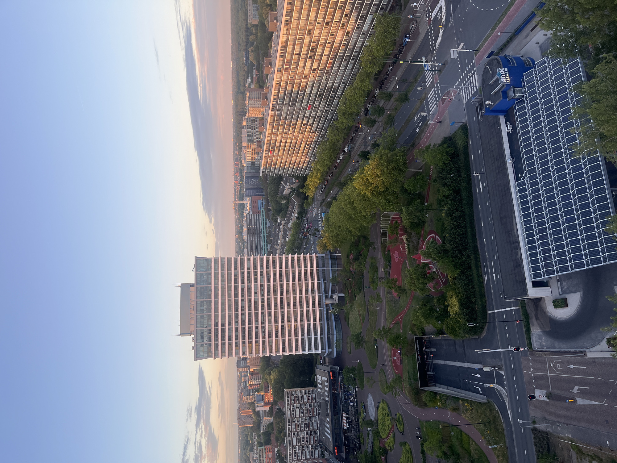 This is a high-angle, long shot of an urban landscape at sunset, likely in the Netherlands, given the architecture and road markings. The sky displays a gradient of soft blues and pinks. In the foreground, a multi-story building with a blue facade and a large solar panel array on its roof is visible. Adjacent to it is a parking garage entrance labeled "Q-PARK BOOTSCHAPSPLEIN."
To the left of the parking garage, a modern building with a white facade and numerous horizontal windows dominates the mid-ground. Its upper section features a more angular design. In front of this building is a park-like area with red-fenced recreational spaces, trees, and curved walkways.
To the right of the modern building, a long, multi-story residential building stretches across the frame. It has a facade characterized by repeating balconies and windows, with the setting sun casting a warm glow on its surface. A tree-lined street with parked cars runs along its base. In the distance, a dense urban skyline with various buildings and a radio tower is visible, receding into the horizon. The scene captures a typical city environment with a mix of residential, commercial, and recreational spaces, illuminated by the warm light of the setting sun.