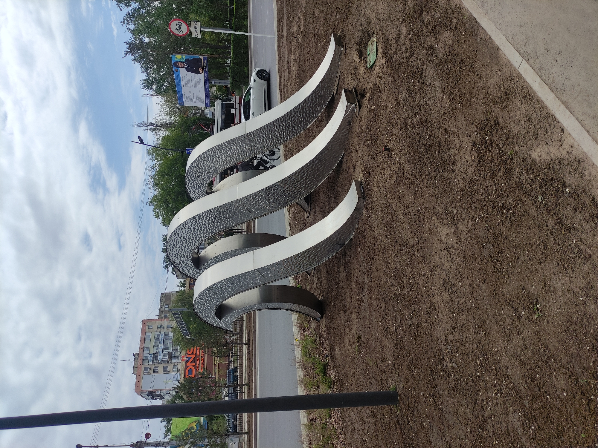 The image shows a three-dimensional, winding, metallic sculpture located in Pavlodar, Kazakhstan. The sculpture consists of three parallel, curved elements with textured inner surfaces and smooth outer surfaces. It is situated in a bare earth area, next to a sidewalk and a road with passing vehicles. A billboard and street signs are visible in the background, along with buildings in the distance. The sky is partly cloudy. A black post is visible in the foreground.