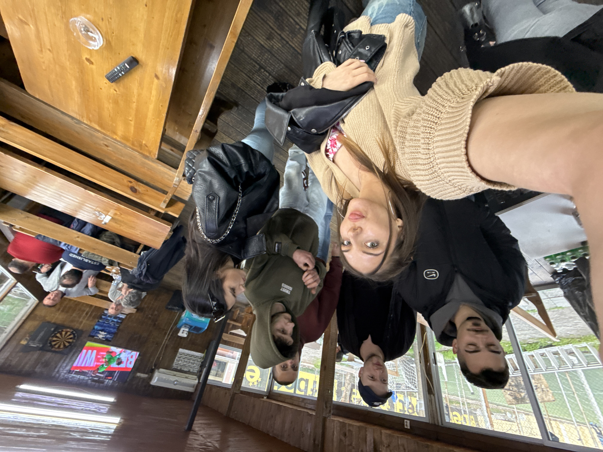 A group of six young adults are posing closely together for a selfie in what appears to be an indoor recreational area, likely a pub or restaurant.
People:
* Six individuals: three women and three men.
* Clothing: One woman is wearing a tan sweater with a visible floral pattern underneath. Another woman is wearing a black leather jacket. One man is wearing a dark hoodie and a cap. Another man is wearing a black Nike jacket over a grey hoodie.
* All appear to be facing the camera.
Environment:
* The setting is a wood-paneled recreational space.
* A wooden table with benches is visible. A remote control and a clear plastic object are on the table.
* In the background, a dartboard and a television screen are visible.
* Outside the windows there is what looks like a green turf field with fencing.
Action/Interaction:
* The group is closely positioned for a selfie, suggesting a social gathering.
Location:
* While it's difficult to confirm with certainty, the presence of a green turf field with fencing visible through the window suggests that the location is in the city of Sofia, Bulgaria.