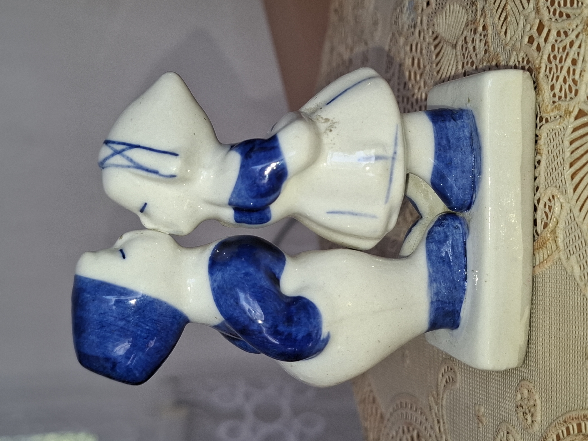 A close-up shot shows a ceramic figurine of two children, a boy and a girl, kissing. The figurine is primarily white with blue accents. The boy is on the left, wearing a blue cap and a blue sash. The girl on the right wears a blue band on her head and a blue sash around her dress. Both figures have their eyes closed. They stand on a white square base. The figurine is placed on a beige, lace-like tablecloth. The background is softly blurred, with hints of light and shadow suggesting an indoor setting. The image does not provide any information about the city of Almaty, Kazakhstan, or any specific location within it.