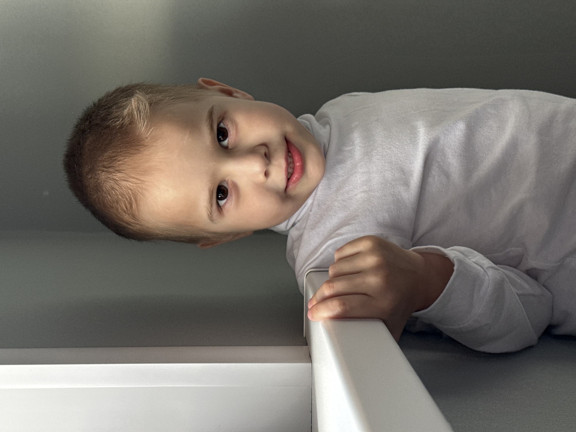 A young boy in a long-sleeved white shirt is looking at the camera. He is holding onto a white structure, possibly a bunk bed frame. The background is a light gray wall and ceiling. There is no verifiable information to confirm this is in Chaikovsky, Russia.