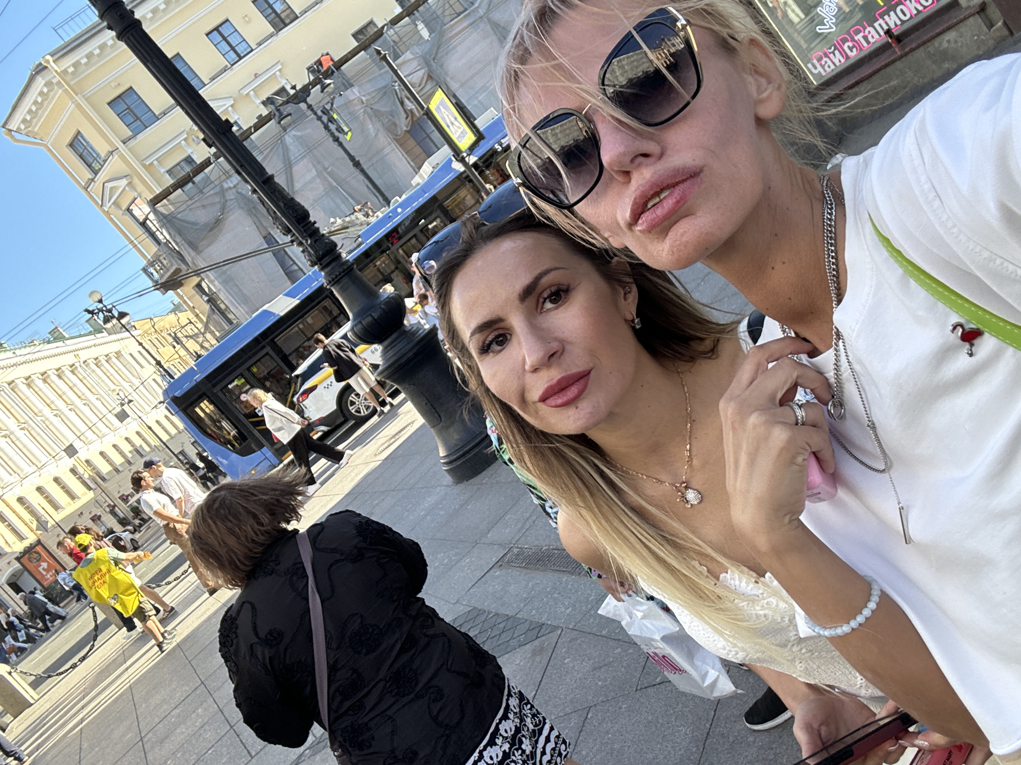 The media file is a selfie taken on a sunny day in Saint Petersburg, Russia, likely during daylight hours given the bright sunlight and clear blue sky. The foreground features two women posing for the camera. The woman on the right has blonde hair, is wearing large sunglasses, a white t-shirt, and multiple necklaces. The woman on the left has long brown hair, is wearing a light-colored top, and has earrings and a necklace. Both women are smiling or have neutral expressions, suggesting a casual and pleasant outing.
In the background, a blue tram is visible, indicating a street scene with public transportation. Several other people are walking on the sidewalk and crossing the street. Buildings with a distinct architectural style, characteristic of Saint Petersburg, line the street. One building appears to be under renovation, covered with a large tarp.
Notable details include the presence of a lamp post and a black metal railing in the foreground, which partially obscures the view of the background. A person in a black jacket and patterned skirt is seen from behind in the lower left quadrant, and another person in a yellow vest is visible further back, potentially a street vendor or guide. A shopping bag with some text is held by the woman on the right. There is a small decorative pin on her t-shirt that appears to be a flamingo. Some text is visible on signs in the background, but it is not clear enough to be transcribed. The overall atmosphere is that of a typical urban day in a city with historical architecture and active street life.