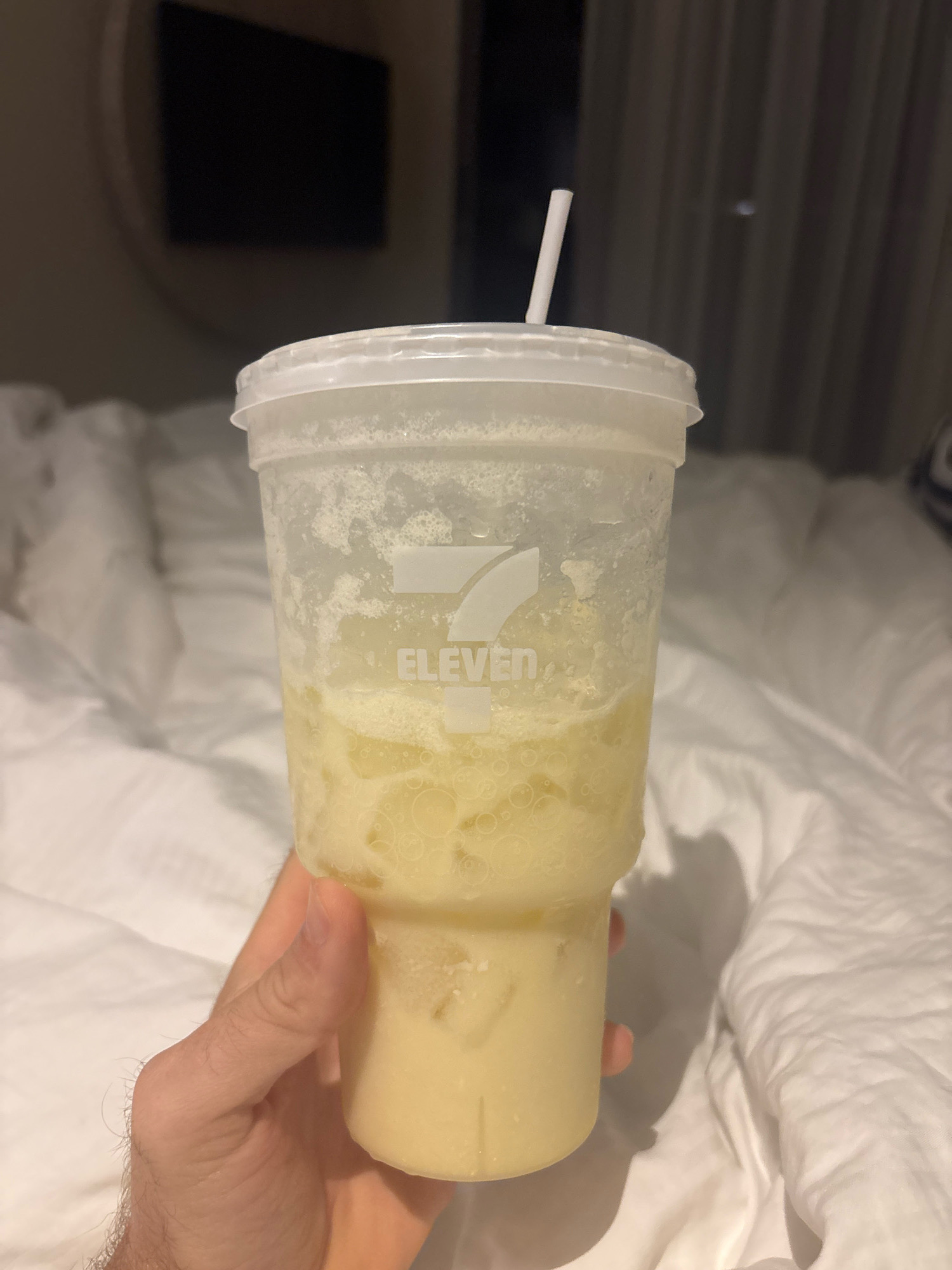 In Karon, Thailand, a hand holds a 7-Eleven branded plastic cup filled with a frothy, pale yellow beverage. The cup has a white lid with a white straw inserted. The beverage appears to contain ice and possibly fruit pulp. The background is blurred, showing what looks like a bed with white sheets and a dark curtain. A dark, rectangular object, possibly a television screen, is visible in the upper left portion of the background. The overall lighting suggests an indoor setting, possibly at night.