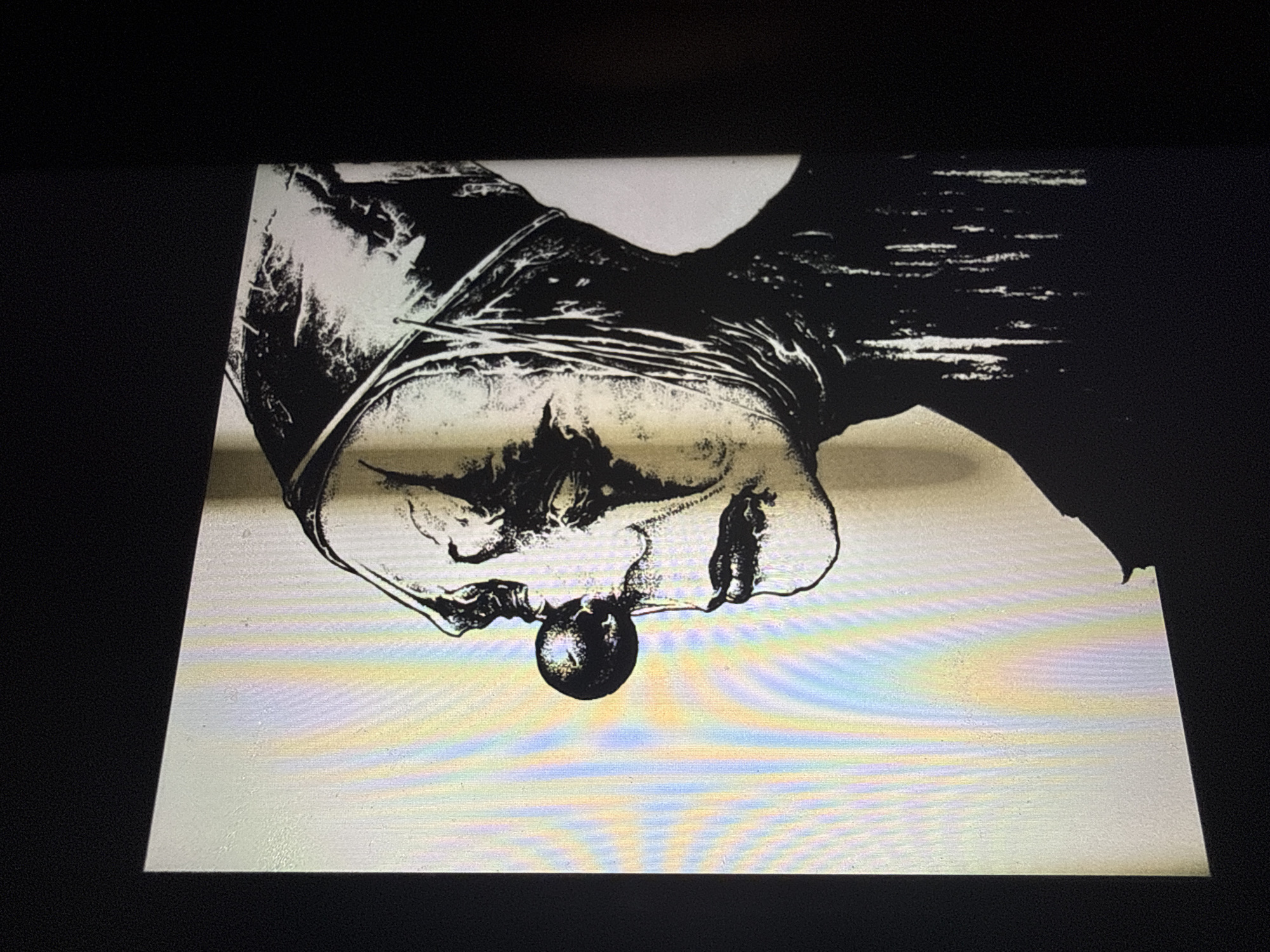 The image is a close-up, high-contrast black and white illustration of a clown's face in profile. The clown wears a conical hat and has a red sphere for a nose. The face is gaunt with deep-set eyes and a sad expression. The background is a pale yellow with vertical rainbow-colored streaks. The image is presented on a screen, as indicated by the dark borders around it and the visible pixels. There is no indication of the location in the city of Sofia, Bulgaria.
