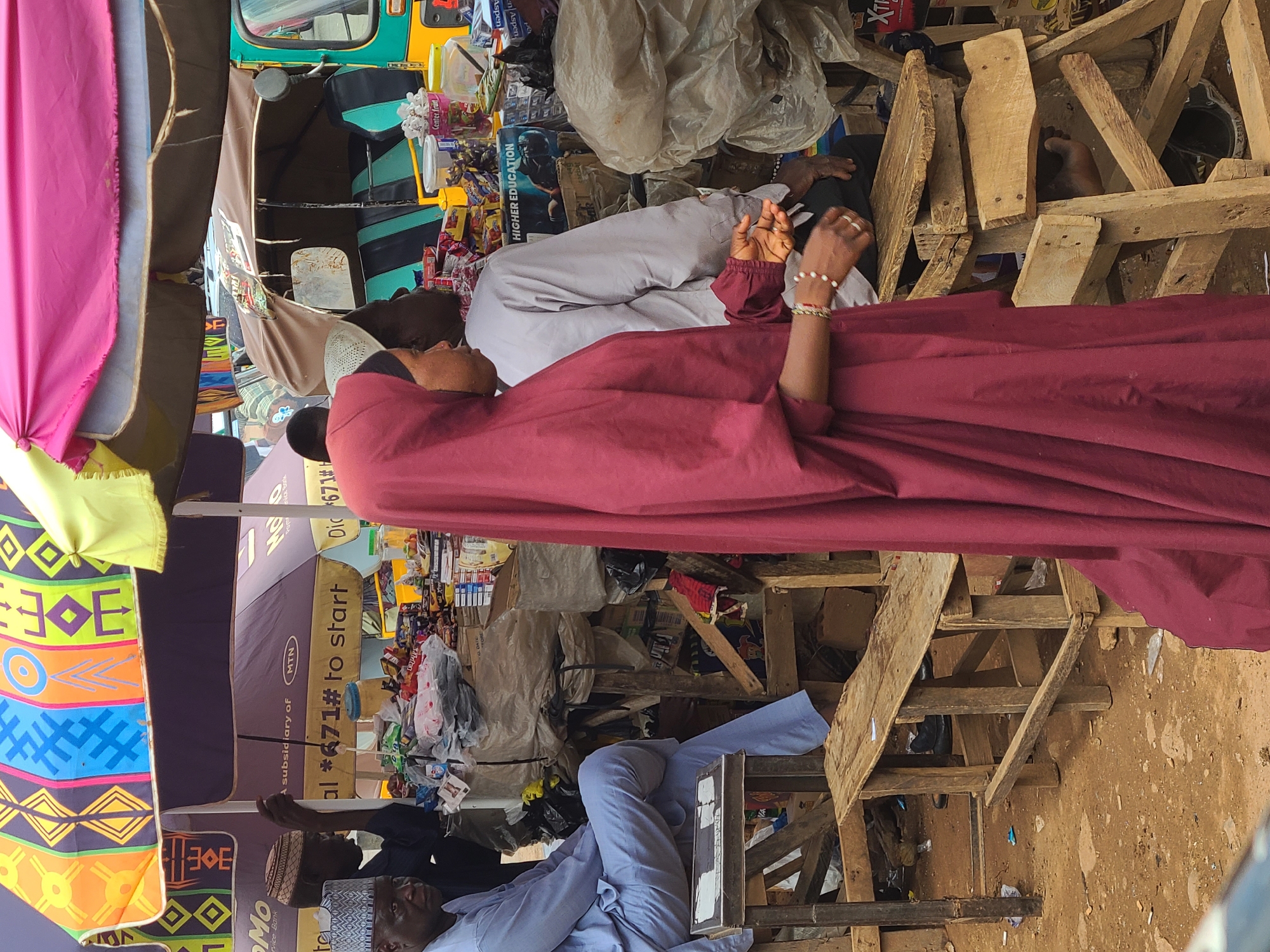 This image captures a vibrant and somewhat chaotic outdoor market scene in Gura Top, Nigeria, filled with people, goods, and makeshift structures under bright daylight.
**Subjects:**
Several individuals are visible. In the foreground, a person in a maroon-colored abaya-like garment and matching head covering stands or sits, with hands adorned with rings and beaded bracelets. Behind them, another person is seen resting, wearing a light grey garment and a white kufi (cap). Towards the bottom, two men wearing traditional caps are present, one in light blue attire, seemingly resting on a wooden structure, and another in a darker shirt. A bare foot is visible tucked among some wooden planks, hinting at another resting individual.
**Objects:**
The scene is densely packed with objects. A green and yellow Keke Napep (auto-rickshaw) is visible towards the top. Market stalls are constructed from rough wooden planks, holding various packaged goods and products. Colorful fabrics (pink, yellow, patterned with geometric designs) hang on the left, possibly for sale or as canopy decorations. Numerous large umbrellas, some plain dark (brown/purple) and others brightly patterned, provide shade; several bear the "MTN" logo. Clutter, including plastic bags, cardboard boxes, and assorted merchandise, fills the spaces between people and structures.
**Activity or event taking place:**
The image depicts a typical day at an open-air market or street side, characterized by commerce, waiting, and resting. People are either engaged in selling/buying or taking a break from the market's hustle.
**Notable details:**
The time of day is clearly daytime, indicated by the bright, natural lighting. The use of umbrellas suggests sunny weather. The ground is dusty dirt, typical of an outdoor market in this region. The Keke Napep and traditional attire provide strong cultural context for Nigeria. The overall atmosphere is busy and functional, reflecting daily life.
**Visible text:**
Text elements include "Center fruit," "HIGHER EDUCATION," "Aspen," and "XTE" on various product packaging. Prominently displayed on umbrellas are "MTN" logos and phrases like "A subsidiary of MTN" and "Dial *671# to start," which likely refers to a mobile network service. Other partially visible texts include "Mo" (possibly part of "Omo Service Bank") and "rvice Bank."