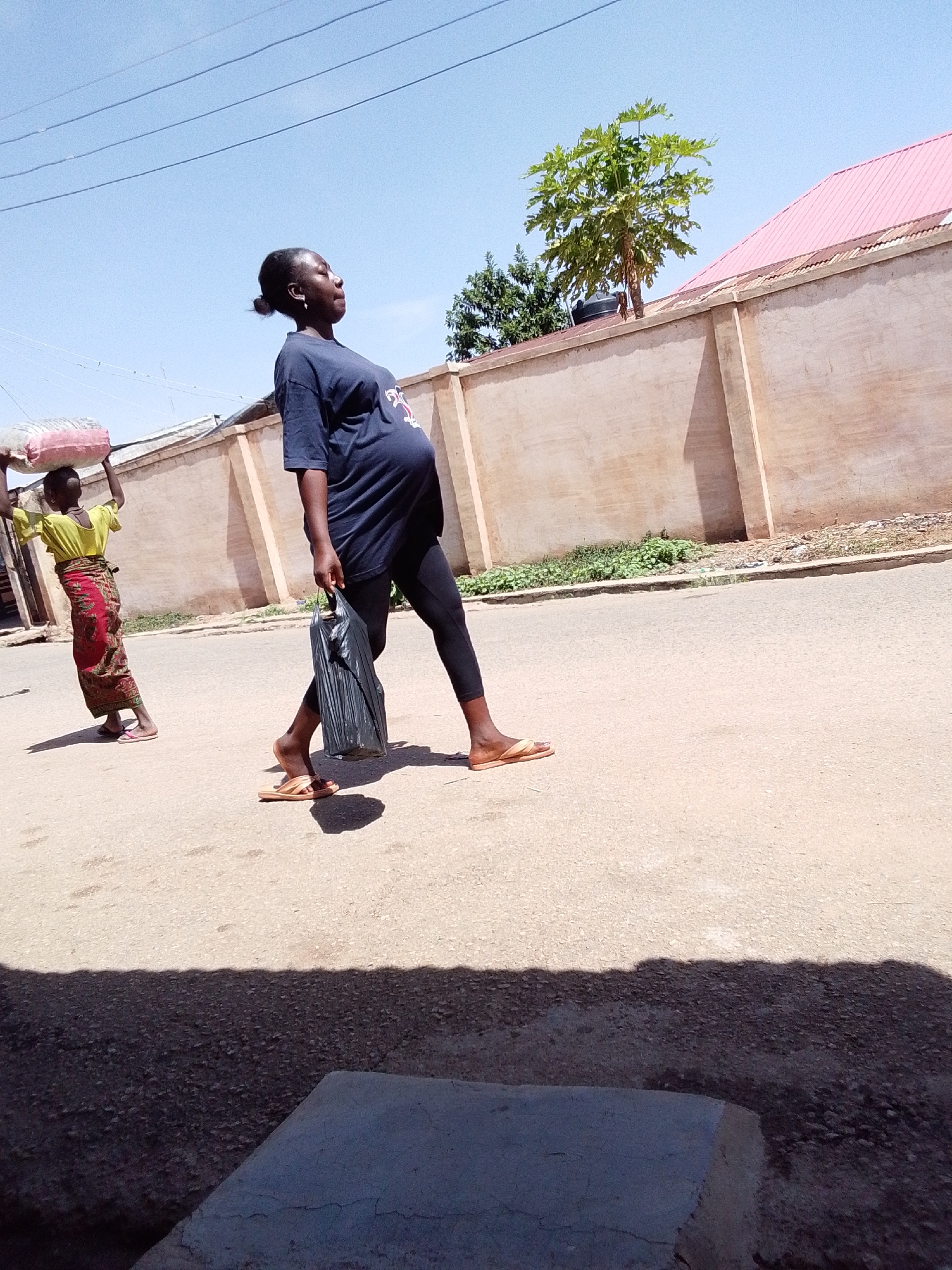 The image depicts a bright, sunny daytime scene on a street in Jos, Nigeria. In the foreground and center, a pregnant woman is walking on a paved road, moving towards the right side of the frame. She wears a dark blue t-shirt that accentuates her pregnancy, black leggings, and light brown slip-on sandals. She carries a black plastic bag in her left hand. Her hair is pulled back, and her head is tilted slightly upwards, suggesting she might be looking ahead or shielding her eyes from the sun.
Further in the background, to the left of the pregnant woman, another individual, possibly a young person, walks in the same direction. This person is carrying a large, light-pink sack balanced on their head and is dressed in a yellow short-sleeved top and a patterned red and green skirt, along with light-colored flip-flops.
The setting includes a long, light-colored concrete wall running along the right side of the road. Behind this wall, a house with a distinct pink corrugated roof and green trees are visible against a clear blue sky. Faint utility lines stretch across the upper part of the sky. The strong shadows cast by the subjects and the wall indicate a bright, high sun, likely midday or early afternoon. A concrete block or curb is partially visible in the shadowed bottom-left corner of the foreground. The scene captures an everyday activity in a warm, outdoor urban or semi-urban environment. No discernible text is visible on any objects or clothing.