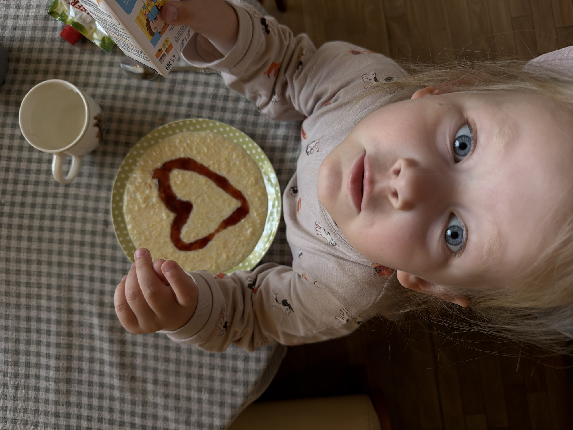 A top-down photographic depiction of a young child seated at a table in Khimki, Russia. The child, with blonde hair and blue eyes, is looking directly upward, engaging with the camera. The child is wearing a light-colored long-sleeved garment patterned with small animal motifs. The child's left hand is visible, holding a small white carton displaying colorful illustrations and an "EAC" conformity mark. On the table, covered by a green and white checkered tablecloth, a light-colored bowl containing porridge is present, featuring a heart-shaped design made from a reddish-brown liquid. The bowl has a light green rim with white polka dots. Adjacent to the bowl, an empty white ceramic mug with a handle and a partial design is visible, along with a silver spoon. A small green and white food pouch with a red cap is also present on the table. The immediate background shows a wooden floor.