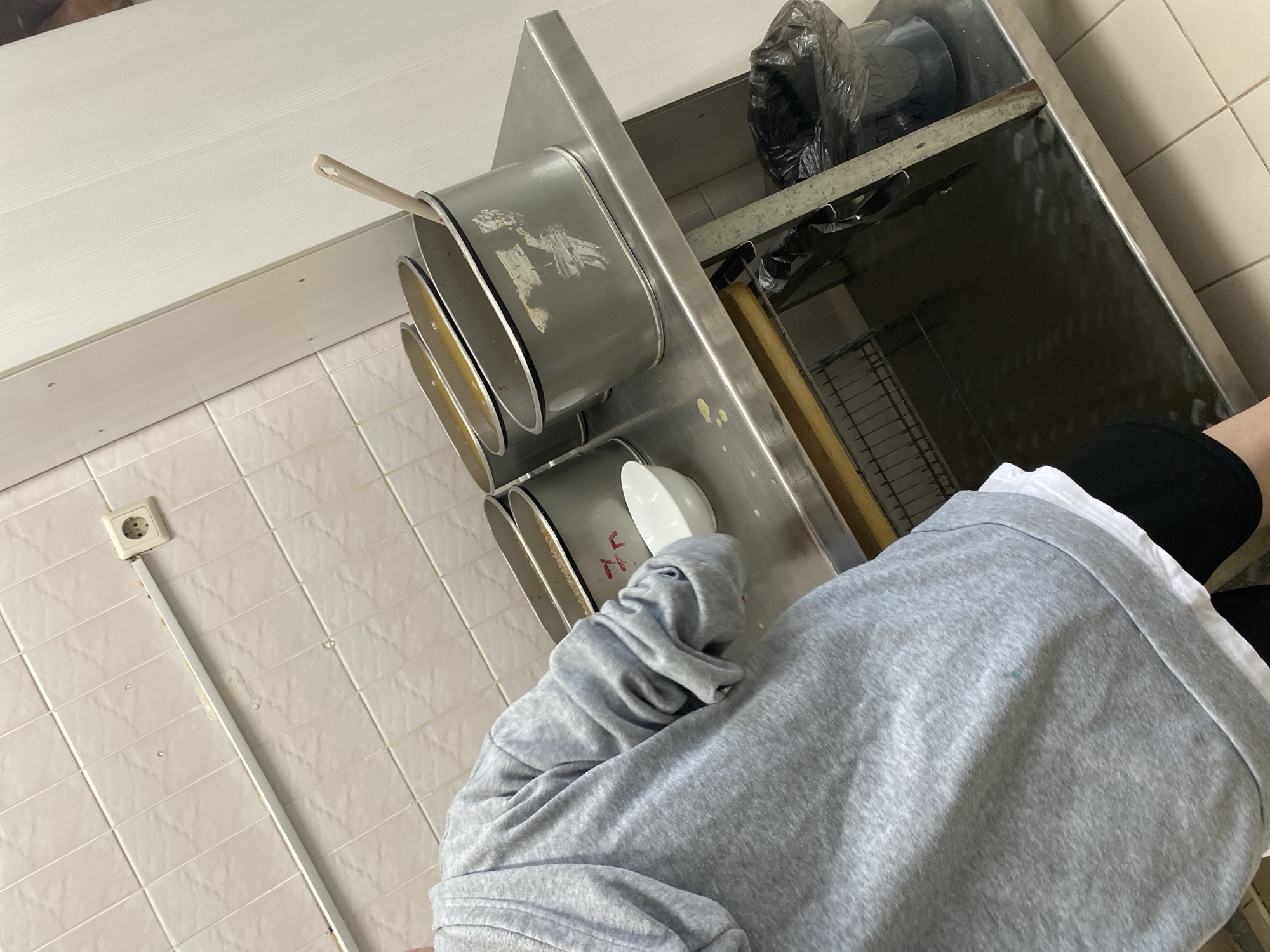 A medium, angled shot captures a scene in a commercial kitchen setting. The camera angle is from above, looking down towards a person standing at a stainless-steel workstation.
The person is wearing a gray hooded sweatshirt over a white undershirt and black pants. Their face is obscured by the hood of the sweatshirt.
The stainless-steel workstation has several rectangular food containers mounted in a staggered configuration, and some contain food. A white serving spoon rests in one of the containers. A black garbage bag is visible on a shelf within the workstation. The workstation appears to be positioned against a white wall.
The floor is tiled with light-colored square tiles. An electrical outlet is visible on the wall.
Based on the presence of Russian text on the garbage bag, the location is presumed to be in the city of Марковский, Russia.