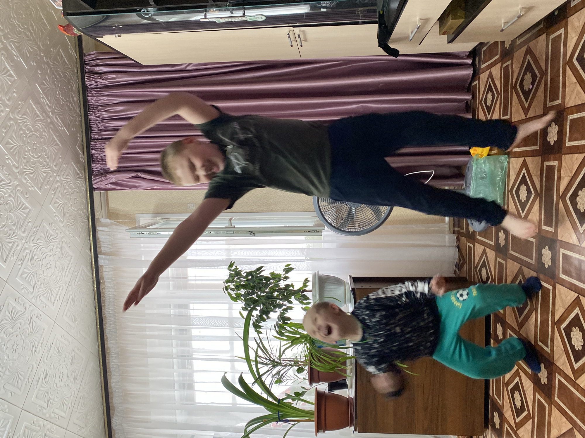 A photo depicts two boys indoors. One boy, in a green t-shirt and dark pants, is leaping in the air. The other boy, wearing a black and white patterned shirt and green pants, appears to be standing on the floor. In the background, there are sheer curtains, a potted plant, a fan, a curtain, a TV, and a tiled floor. The photo was taken in a place that has been identified as Bely Klyuch, Russia.