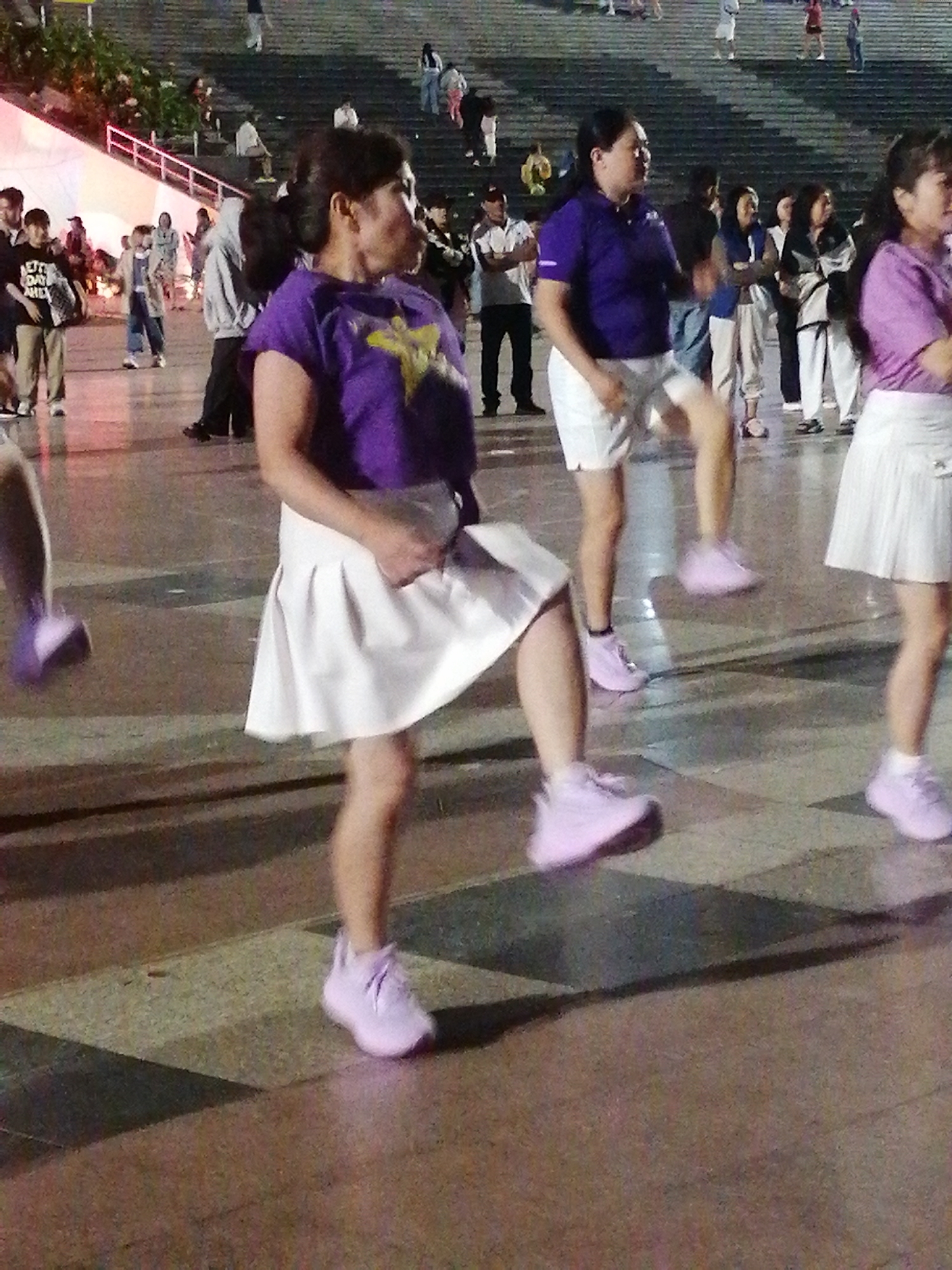 This media file captures a group of people, primarily women, engaged in an outdoor exercise activity, likely a dance or aerobic session. They are dressed in activewear, with several individuals wearing purple tops and white skirts or shorts, and light pink sneakers. The scene appears to be an urban public space with a tiled floor, suggesting a plaza or paved area. In the background, tiered seating or stairs are visible, occupied by other spectators or participants. The lighting suggests it is evening or late afternoon, with artificial lights illuminating the area.
The primary activity is a synchronized movement exercise, with the individuals in the foreground focusing on their steps and poses. The overall mood seems energetic and communal, with people participating together in a structured activity. The backdrop shows a crowd of onlookers and participants, indicating a public event or gathering. Some of the people in the background are dressed in casual wear, while others are also in athletic attire. There are some decorative elements in the background, including what looks like a stage or elevated platform with lights.
The setting appears to be a well-lit public square, possibly a recreational area or a venue for outdoor events. The presence of the tiered seating suggests this location is designed to accommodate audiences. The flooring, a pattern of dark and light tiles, is a prominent feature of the immediate foreground. The lighting, shadows, and the attire of the participants point towards an organized outdoor event taking place in the evening. There is no visible text in the image.