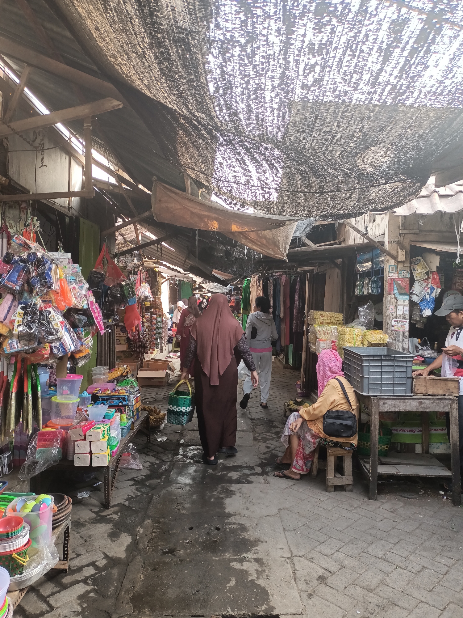 The image depicts a bustling market scene in Badas, Indonesia. The market is characterized by narrow aisles covered by a textured, dark canopy that provides shade. Stalls line both sides of the aisle, displaying a variety of goods, including toys, plastic containers, and clothing. Several individuals are visible, engaged in shopping or working at the stalls. In the foreground, a person wearing a brown hijab and dress is walking away from the camera, carrying a green basket. To their left, shelves are filled with colorful plastic items. Further down the aisle, a woman in a pink hijab is seated on a stool, looking towards the right. Other people are seen browsing at stalls or walking in the background. The ground appears to be paved with bricks and shows signs of being wet. The overall atmosphere suggests a lively and active marketplace.