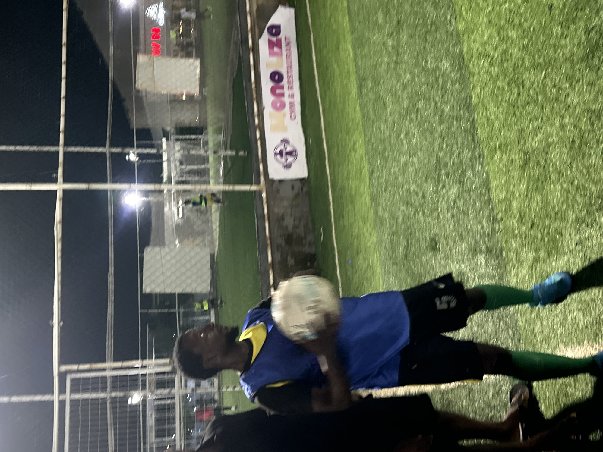 An outdoor, artificial turf soccer field at night in Abuja, Nigeria, is illuminated by bright lights. In the foreground, a Black male wearing a blue vest over a yellow shirt, black shorts with the number '5' on the leg, green socks, and blue soccer cleats, holds a soccer ball in his hands. To his left, another Black male is partially visible. In the background, a white banner with purple text that reads "MONO LIZA GYM & RESTAURANT" is affixed to a concrete barrier. Several other individuals are scattered across the field, some near soccer goals. A building with red illuminated letters that appear to spell "WN" is visible in the distance.