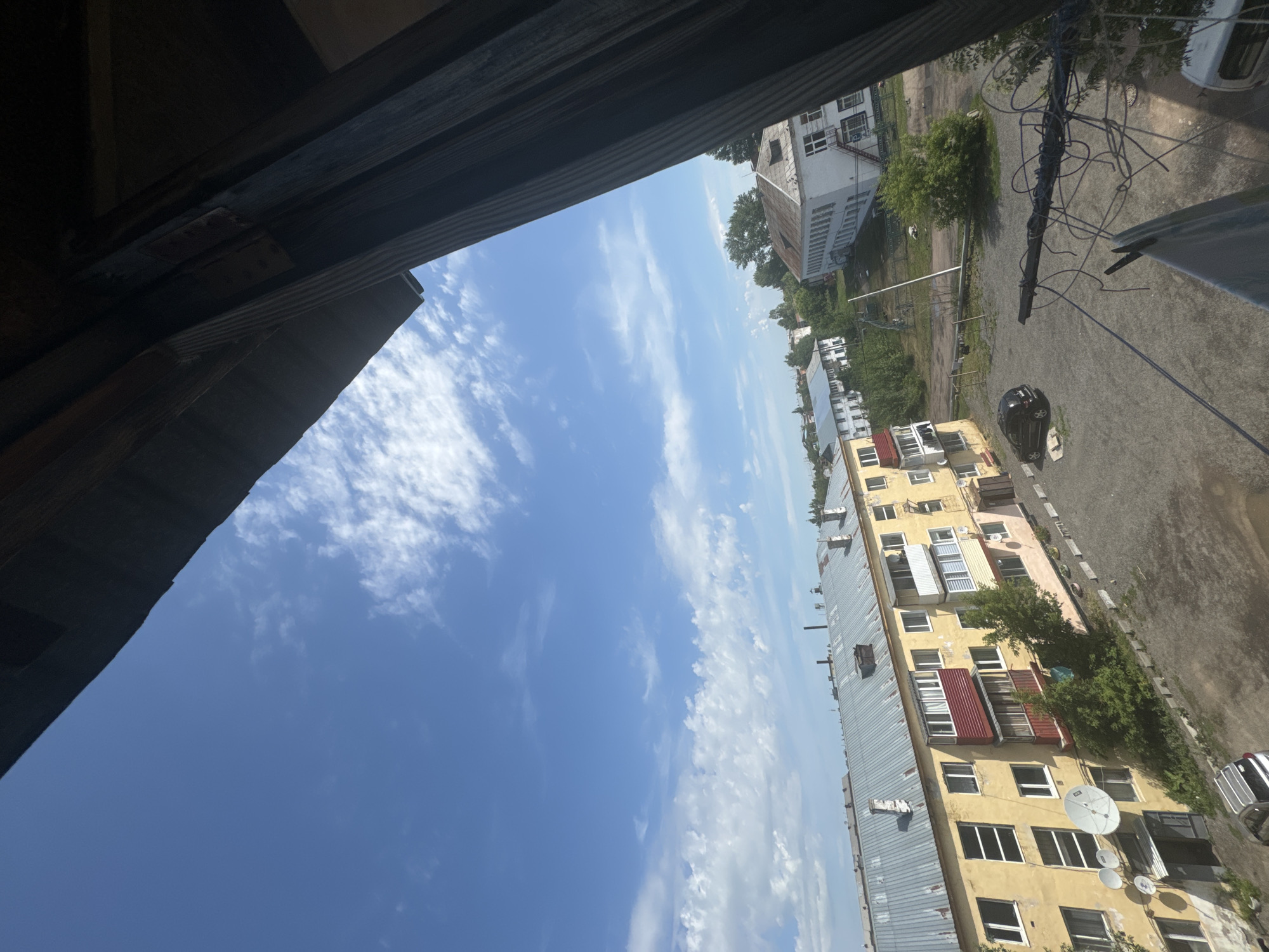 This is an aerial, wide-angle shot of a residential area in the city of Aqköl, Kazakhstan. The scene is dominated by a bright blue sky with scattered white clouds. In the foreground, a dark, wooden structure, possibly a balcony railing or roof overhang, partially obscures the view. Below, a row of Soviet-era apartment buildings is visible. The closest building is yellow with a corrugated metal roof, featuring satellite dishes mounted on its facade. To the right, another building, painted white with a similar metal roof, stands slightly further back. The ground is a gravelly area with some patchy vegetation and a few parked cars, including a dark SUV and a white van. The overall impression is one of a modest, working-class urban environment under a clear, sunny sky.