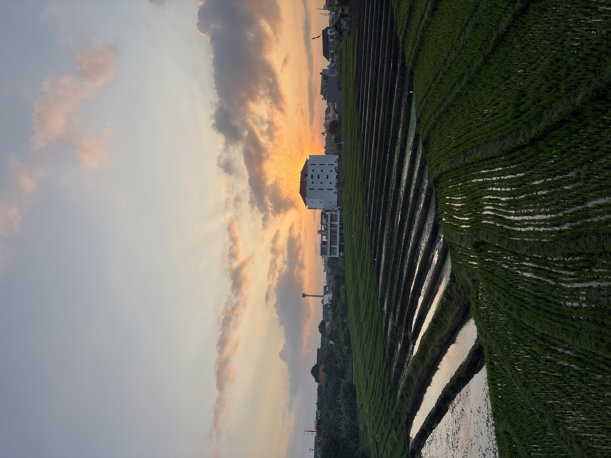 The image depicts a picturesque sunset over terraced rice fields in North Kuta, Indonesia. The sky is a canvas of warm oranges, pinks, and soft blues, with the sun peeking through scattered clouds. In the mid-ground, a modern, multi-story white building stands out against the lush greenery. The rice paddies, some filled with water reflecting the sky, others with neatly planted green shoots, cascade down the landscape, creating a visually striking pattern. In the distance, other buildings and structures, including a tall communications tower, are visible, hinting at the proximity of a more developed area. The overall scene conveys a sense of tranquility and natural beauty, characteristic of rural Indonesia.