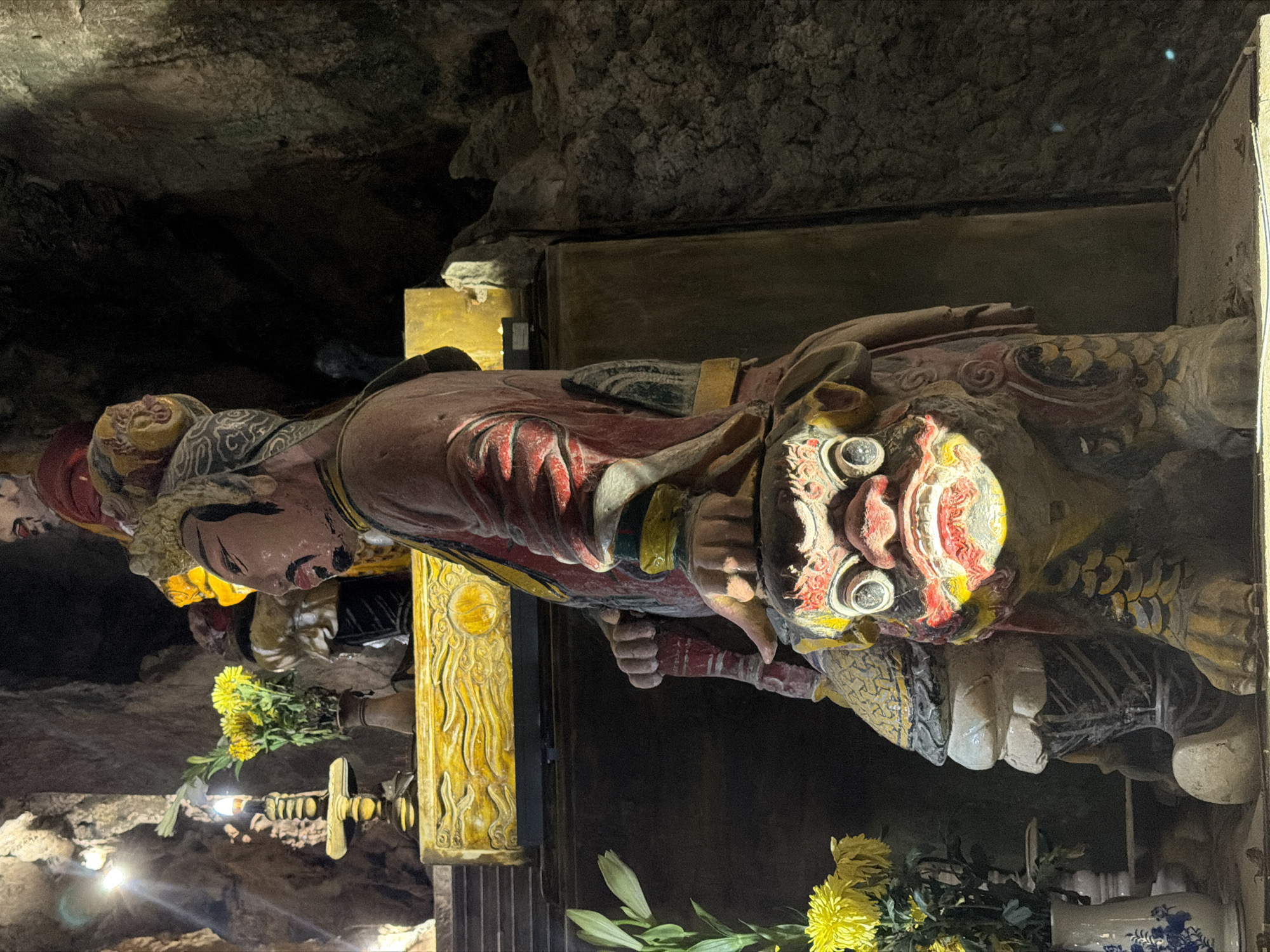 A colored carving of an Asian deity or figure, facing left, is prominently featured in the frame. The carving is detailed with red, yellow, and black markings and features. The head has a mask-like face.
The statue is located in a cave temple in the Marble Mountains, a cluster of limestone and marble hills located in Đà Nẵng, Vietnam. The cave environment has rough, unfinished walls. The area is decorated with yellow chrysanthemum flowers and a column or pillar in the middle of the image.