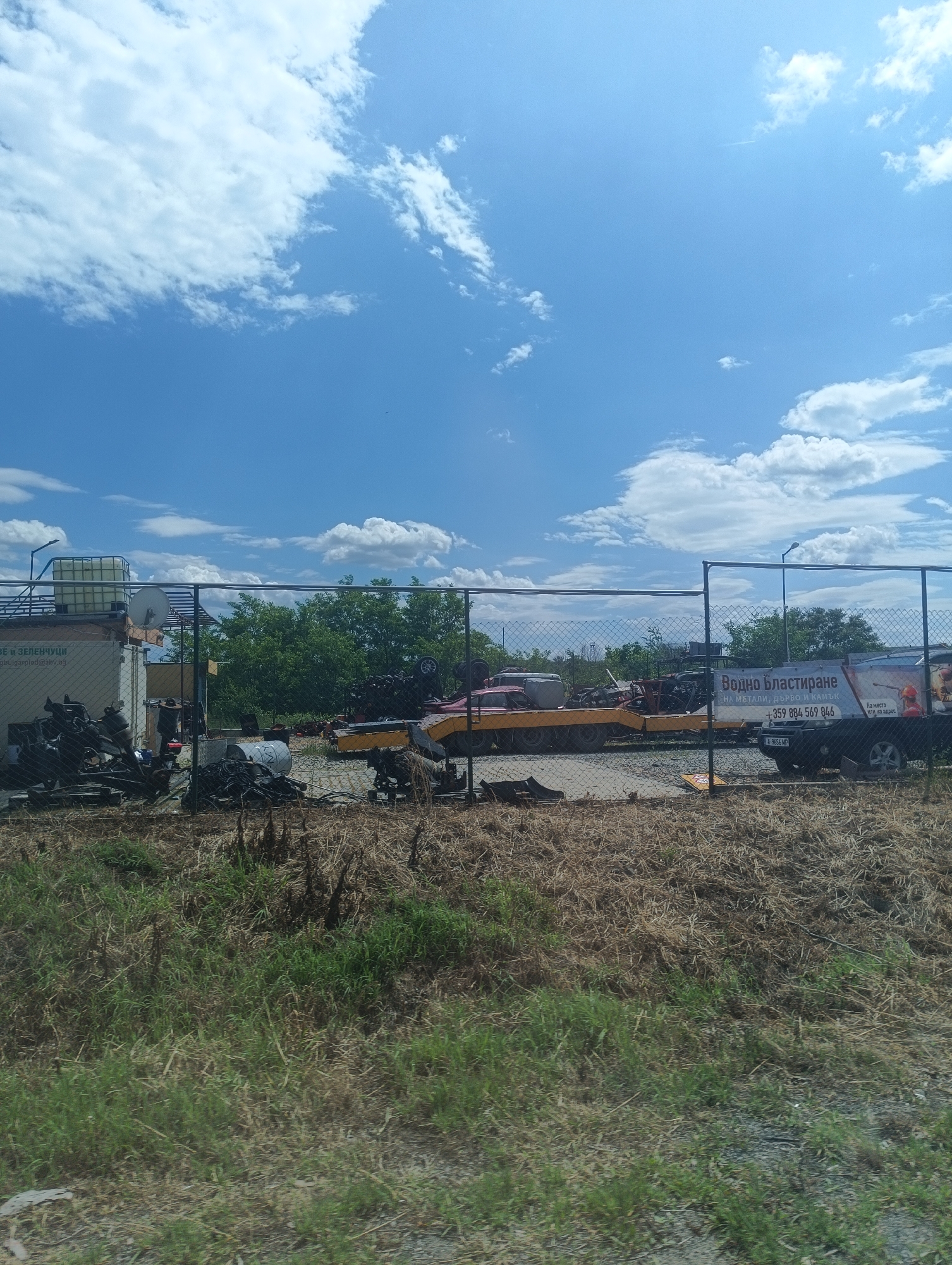The image shows a fenced-off area, likely a junkyard or storage facility, on a sunny day. In the foreground is grass and dry vegetation. Beyond the chain-link fence, there are piles of scrap metal, machinery, and possibly old car parts. In the background, there is a trailer with various items and an advertising sign written in Bulgarian, which translates to "Water blasting - for metals, wood, and stone" along with a phone number (+359 884 569 846). There is a black pickup truck parked next to the trailer. The sign also contains the text "On-site or at an address." Behind this area are green trees and blue sky with clouds. The sign near the white building contains "ВЕ И ЗЕЛЕНЧУЦИ" and "gbulgarpled@abv.bg." The license plate of the vehicle is A 9656 MP. The location is likely in Bulgaria, potentially near Burgas.