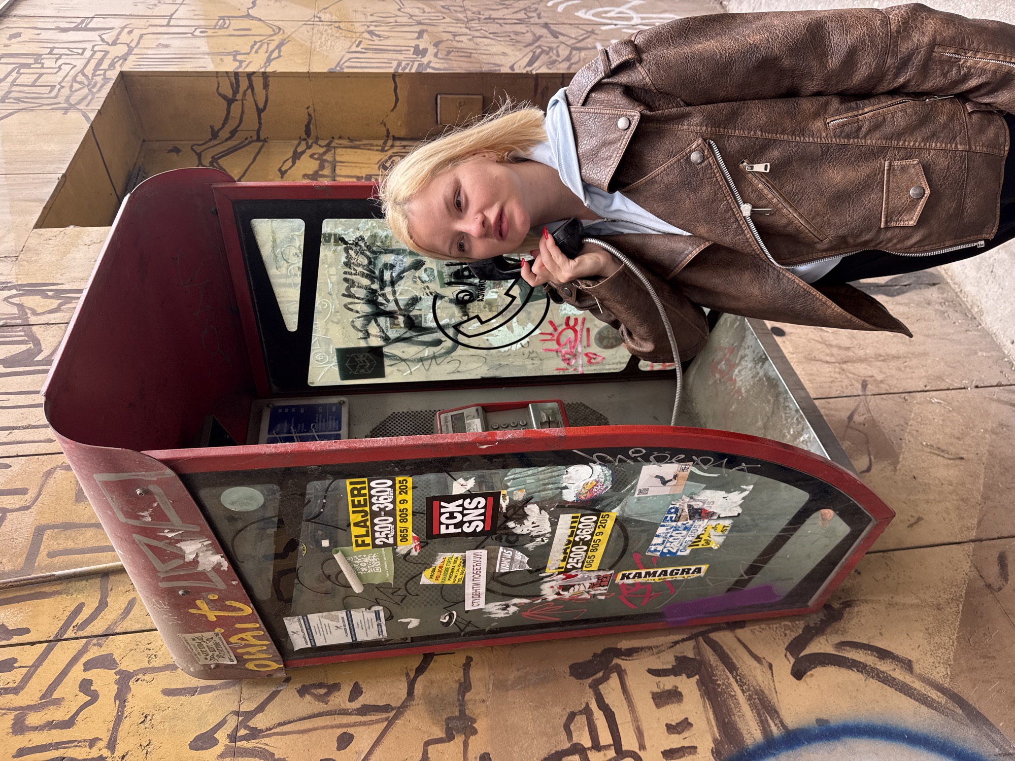 A woman with blond hair is pictured inside a red public telephone booth, holding the receiver to her ear. She is wearing a brown leather jacket over a light blue hoodie. The phone booth is covered in graffiti and stickers, including one that says "FLAJERI 2500-3600 065/805 9 205" and another that says "FCK SNS". The exterior of the phone booth and the wall behind it are also covered in graffiti. The location is in the city of Belgrade, Serbia.
