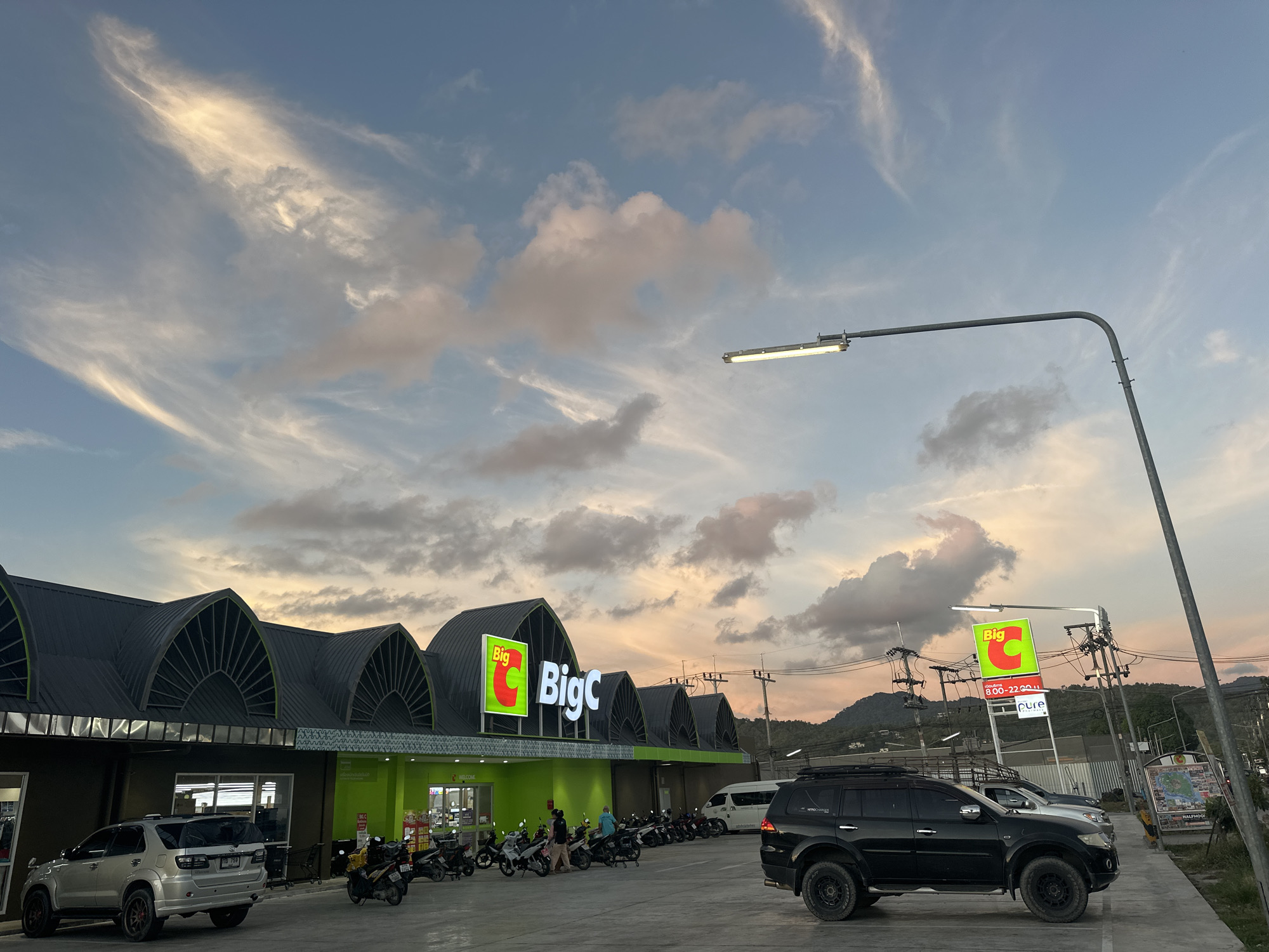 The image shows the exterior of a Big C supermarket in Koh Phangan, Thailand, during a sunset. The sky is filled with wispy clouds tinged with pink and orange hues, indicating the setting sun. The supermarket has a distinctive green facade with large "Big C" signage illuminated in red and white. Several motorcycles are parked in front of the store, along with two cars: a silver SUV on the left and a black SUV on the right. A street lamp with a modern design is visible in the upper right portion of the image, casting light on the parking lot. The overall environment appears to be a commercial area on the island, with some vegetation and hills visible in the background.