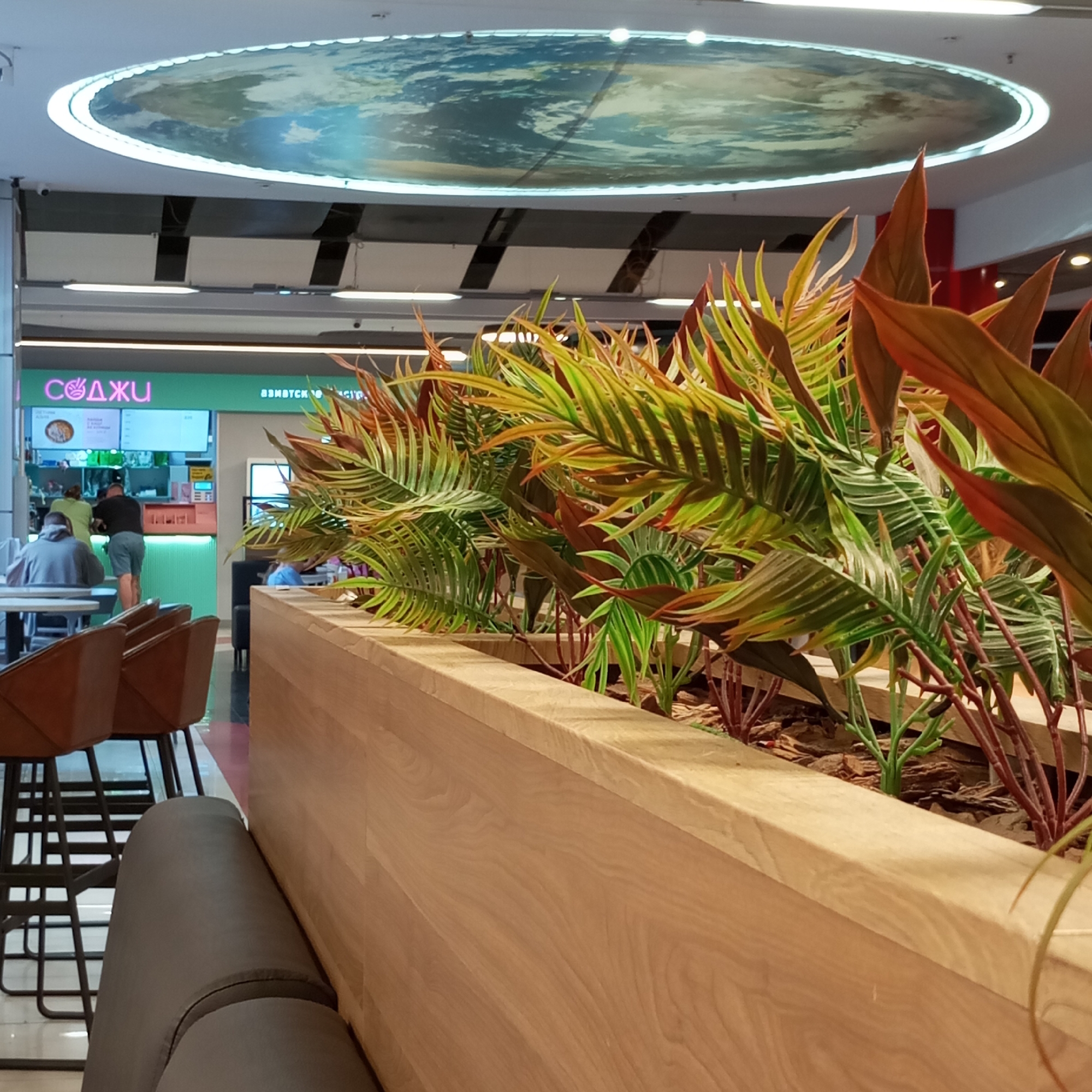 The image depicts the interior of a food court or dining area, likely located in a shopping mall in Izhevsk, Russia, suggested by the signage in Russian. In the foreground, a wooden planter box contains artificial plants with vibrant green and reddish-brown leaves. Behind the planter, a row of brown leather bar stools with black metal frames are visible. To the left, a food stall with a green counter and a menu board is serving customers. Two individuals are interacting with the staff at the counter, while another person is seated at a table in the background. The ceiling features a large circular illuminated mural, possibly depicting the Earth. The overall scene is brightly lit, suggesting an indoor public space.