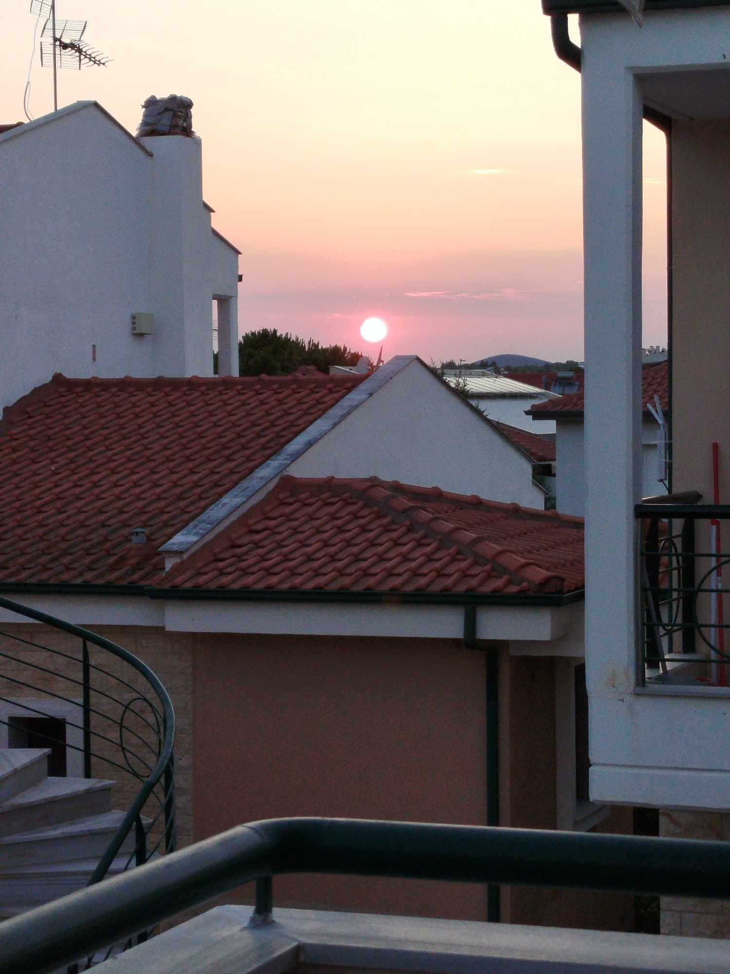 The image captures a sunset view over a residential area in the Sithonia Municipal Unit, Greece. The foreground features a dark metal railing and a portion of a staircase with a spiral metal handrail. Beyond this, terracotta tiled roofs dominate the scene, covering several buildings. A white building with an antenna on its roof is visible on the left, and another white building with architectural details is on the right, obscuring part of the view. In the distance, the setting sun, a bright orange-pink orb, is low in the sky, casting a warm glow over the landscape. Silhouetted trees and hills can be seen against the horizon. The sky is a soft gradient of pink and yellow, with hints of clouds. The overall impression is one of a peaceful, urban evening.