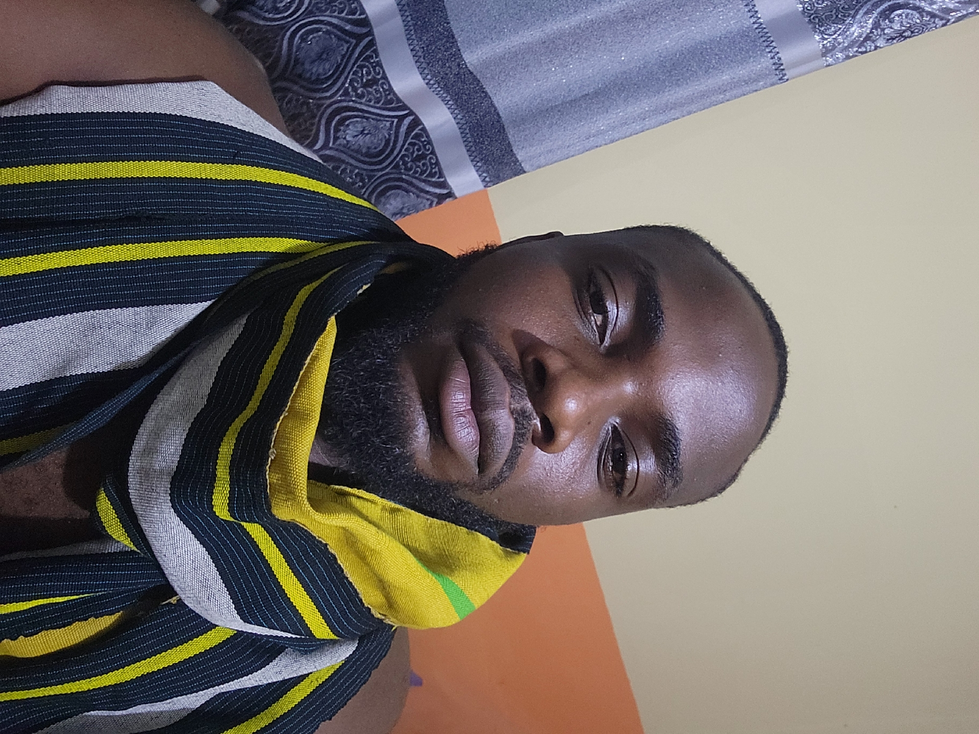The image features a head-and-shoulders portrait of a dark-skinned male individual with a short haircut and a full beard, gazing directly into the camera with a neutral expression. He is draped in a woven garment or scarf displaying vertical stripes in yellow, dark blue (or black), and white. The background consists of a plain light beige wall behind his head, an orange wall segment to his right, and a gray curtain with a repeating ornate dark pattern visible on the left side of the frame. The scene is indoors.