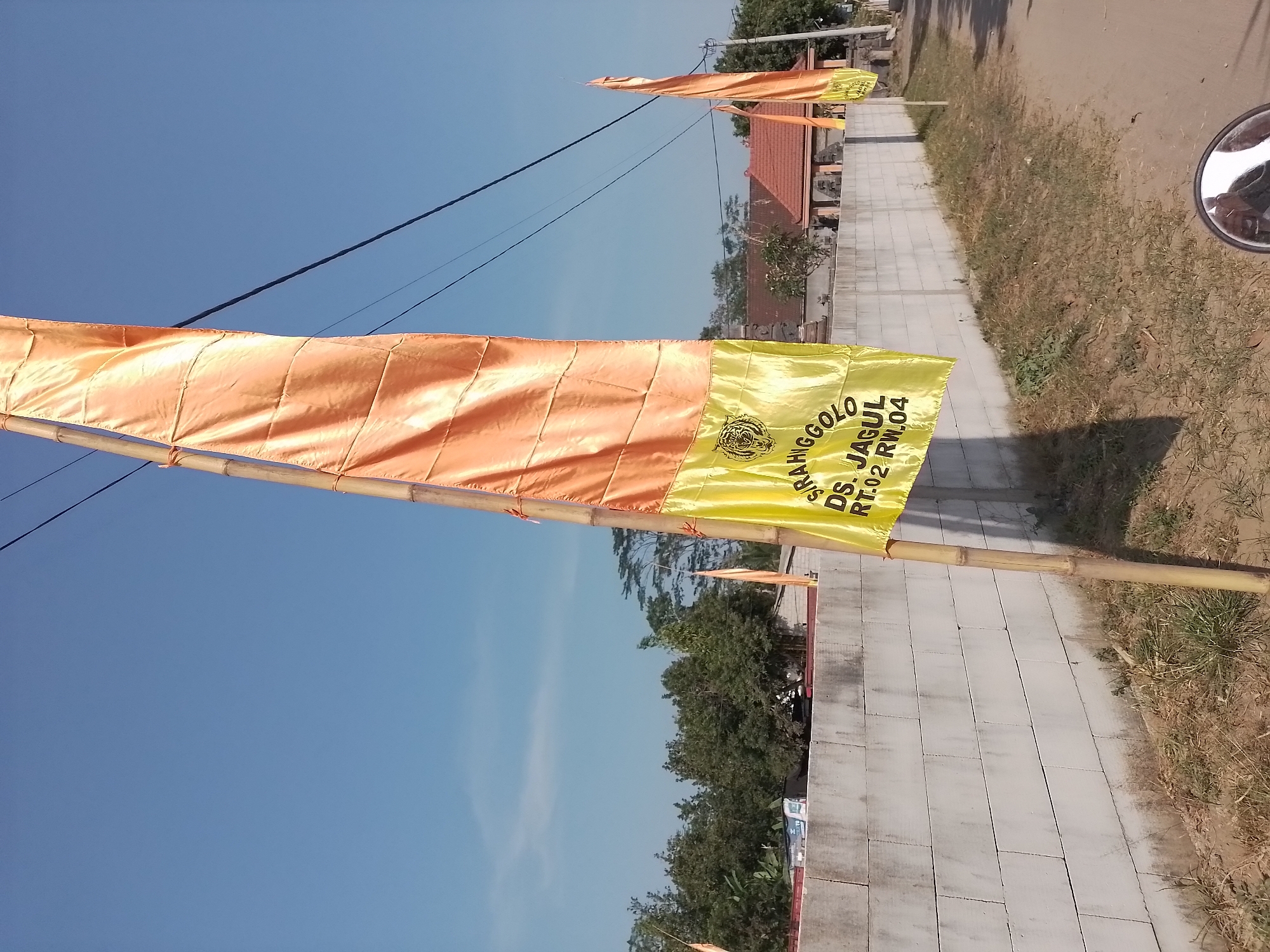 The image depicts an outdoor scene under a clear blue sky in Ngancar, Indonesia. In the foreground, a tall, thin bamboo pole supports a large, orange and yellow flag. The flag is made of a shiny fabric and has text written in black on the yellow section, which appears to read "SIRAHGGOLO DS. JAGUL RT.02 RW.04" and a tiger emblem. To the right of the main flag, another similar orange flag is visible, also on a bamboo pole.
Behind the flags, a long, white concrete block wall stretches horizontally across the frame. Beyond the wall, lush green trees and vegetation are abundant. In the distance, several buildings with terracotta-colored roofs are visible, suggesting a village or residential area. A road runs alongside the wall on the right side of the image. The ground in front of the wall is covered with dry grass and dirt. The perspective suggests the photo was taken from a moving vehicle, possibly a motorcycle, as the reflection of a rearview mirror is visible at the bottom of the frame. The overall impression is a sunny day in a rural Indonesian setting.