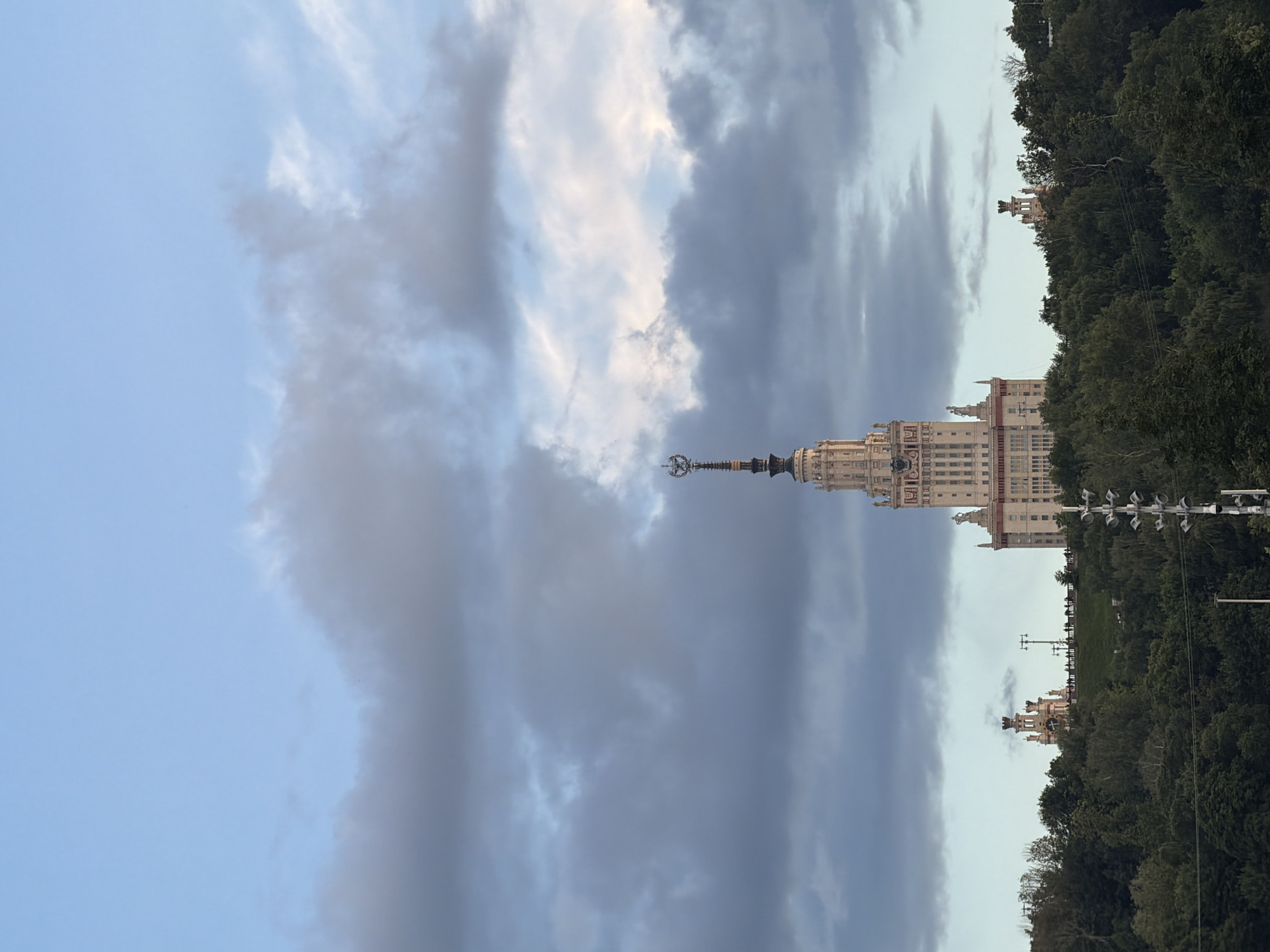 A tall, ornate building with a spire is centered in the image, rising above a dense line of dark green trees. The building is light-colored with many architectural details, including statues and decorative elements. The spire is topped with a star-like ornament. To the left and right of the central building, further back and lower, are two smaller, similarly ornate buildings with spires, also peeking out from behind the trees. The sky above is a pale blue with dramatic, dark gray clouds, some of which are illuminated from behind, creating a soft glow. The overall impression is one of a grand structure set against a moody sky. The location is described as Unknown, Unknown.