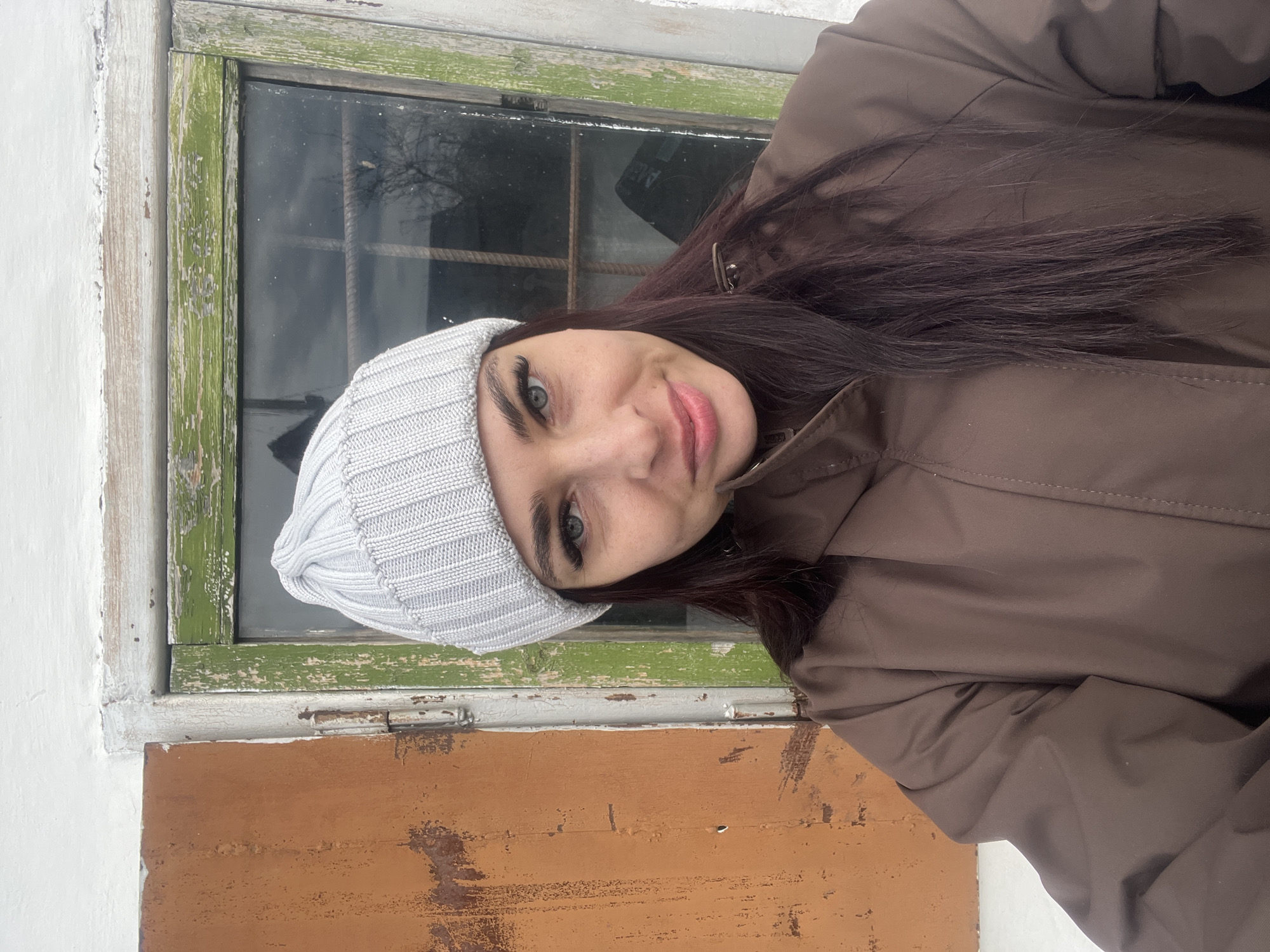 The image shows a woman with long, dark hair and light eyes wearing a light-colored knitted hat and a brown jacket. She is positioned in front of a window with a weathered green frame. Below the window, there's a section with a brown and orange painted surface. The background suggests an outdoor environment. The location is Chaikovsky, Russia.