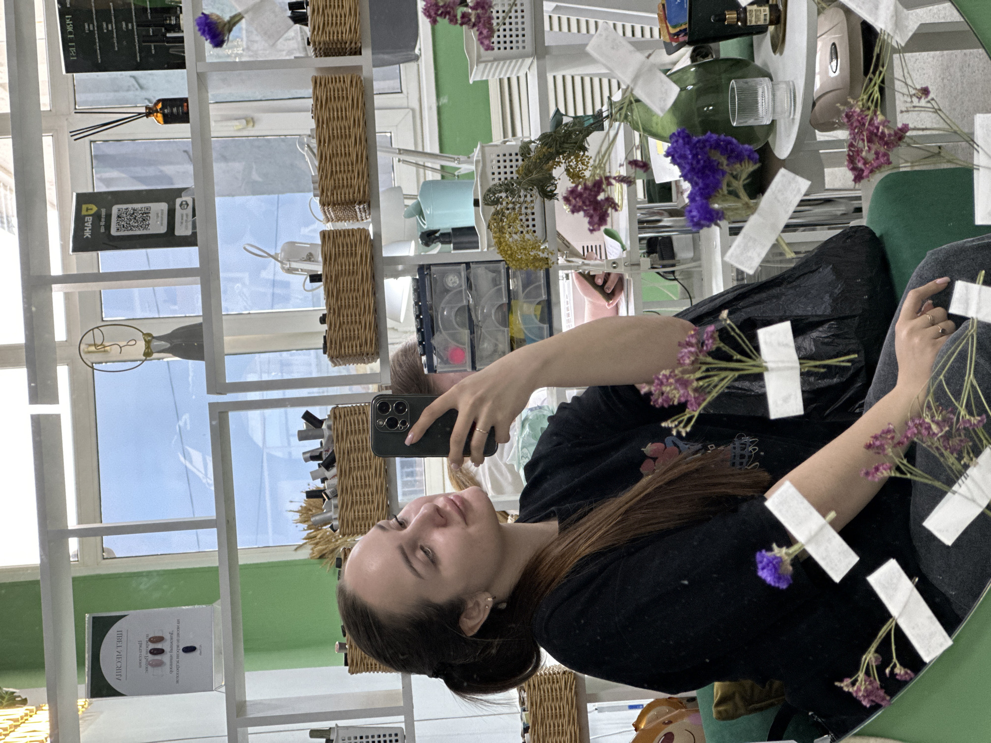 A young woman takes a selfie using a phone in front of a mirror in Chaikovsky, Russia. The mirror is surrounded by shelves holding various items like bottles, baskets, and decorations including dried flowers taped to the glass. The woman is wearing a dark top. The background is filled with a white shelf system and a window letting in daylight.