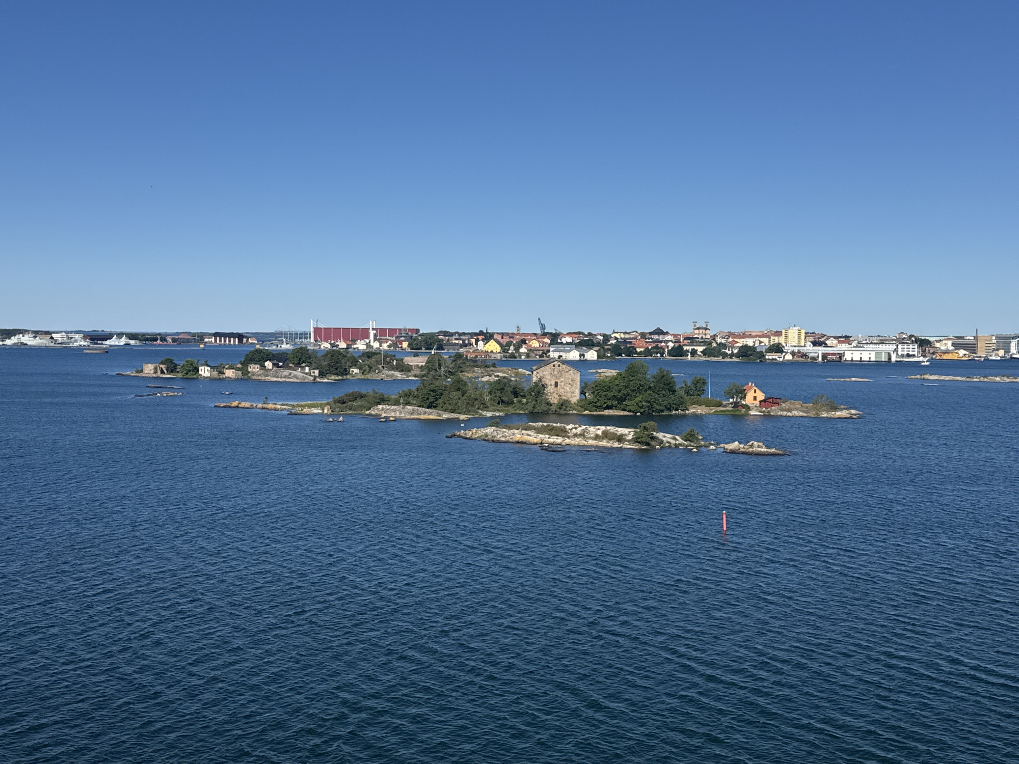 The image shows a view of the archipelago of Karlskrona, Sweden, looking towards the city. The foreground is dominated by a large expanse of dark blue, rippling water. In the middle ground, several small, rocky islands are scattered across the water. Some of these islands are covered in green vegetation, and a few have small buildings or ruins. One island features a prominent stone structure resembling a tower. Further in the distance, the cityscape of Karlskrona is visible, characterized by a collection of buildings with red roofs and some taller structures, including cranes. The sky is a clear, bright blue. Several ships are visible in the water, both near the islands and further out towards the city. A red buoy is present in the water in the right mid-ground.