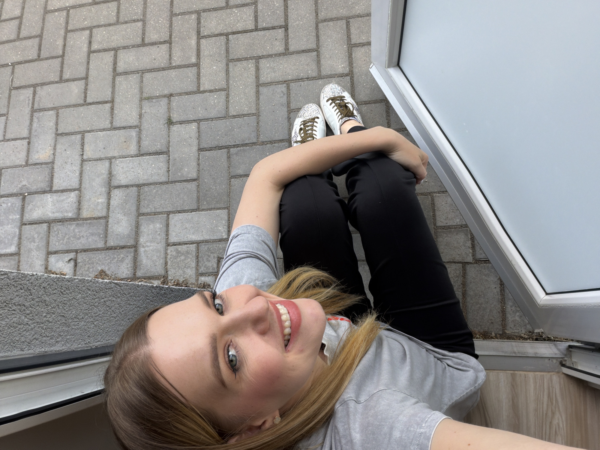 An eye-level, high-angle, slightly tilted medium shot shows a woman with blonde hair, wearing a grey t-shirt and black pants, sitting on the ground outside a building. She is looking up and smiling at the camera. Her left hand is resting on her knee, and her right hand is visible near her thigh. The woman is wearing white sneakers with glittery accents and brown laces. The ground is paved with grey interlocking bricks laid in a herringbone pattern. To the left of the woman is a section of a grey wall and a silver-framed window or door. The image appears to be taken in an urban outdoor setting, possibly a courtyard or sidewalk. There is no specific indication of the location within Riga, Latvia.