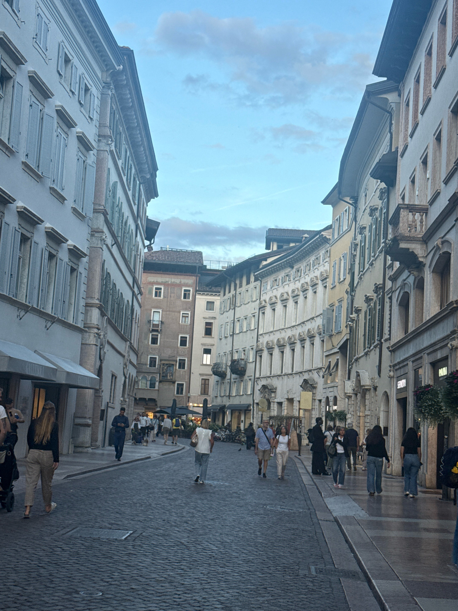 The image depicts a street scene in the city of Trento, Italy, likely during the early evening. The street is paved with cobblestones and is lined with tall, historic buildings featuring arched windows and intricate architectural details. Several people are walking along the street and on the sidewalks.
On the left side of the street, a light-colored building with blue shutters dominates the foreground. A woman with long brown hair, wearing a black top and beige pants, walks away from the camera, pushing a stroller. Another person, partially visible, is to her left.
The street itself curves slightly as it recedes into the distance, flanked by buildings on both sides. The buildings on the right are a mix of architectural styles, with some painted in light colors and others with exposed stone facades. Balconies with ornate railings are visible on some of the buildings.
Several individuals are walking towards the viewer from the distance. One man in a blue shirt and shorts is walking with a woman in white. Further down, a group of people are strolling, some appearing to be browsing shops. A man in a dark shirt and a dark cap walks with his phone in hand.
The sky above is a soft blue with scattered clouds, suggesting the end of the day. The overall atmosphere is that of a bustling European city street with people enjoying an evening stroll. The shops lining the street appear to be open, as indicated by awnings and signage. The lighting is subdued, hinting at twilight.