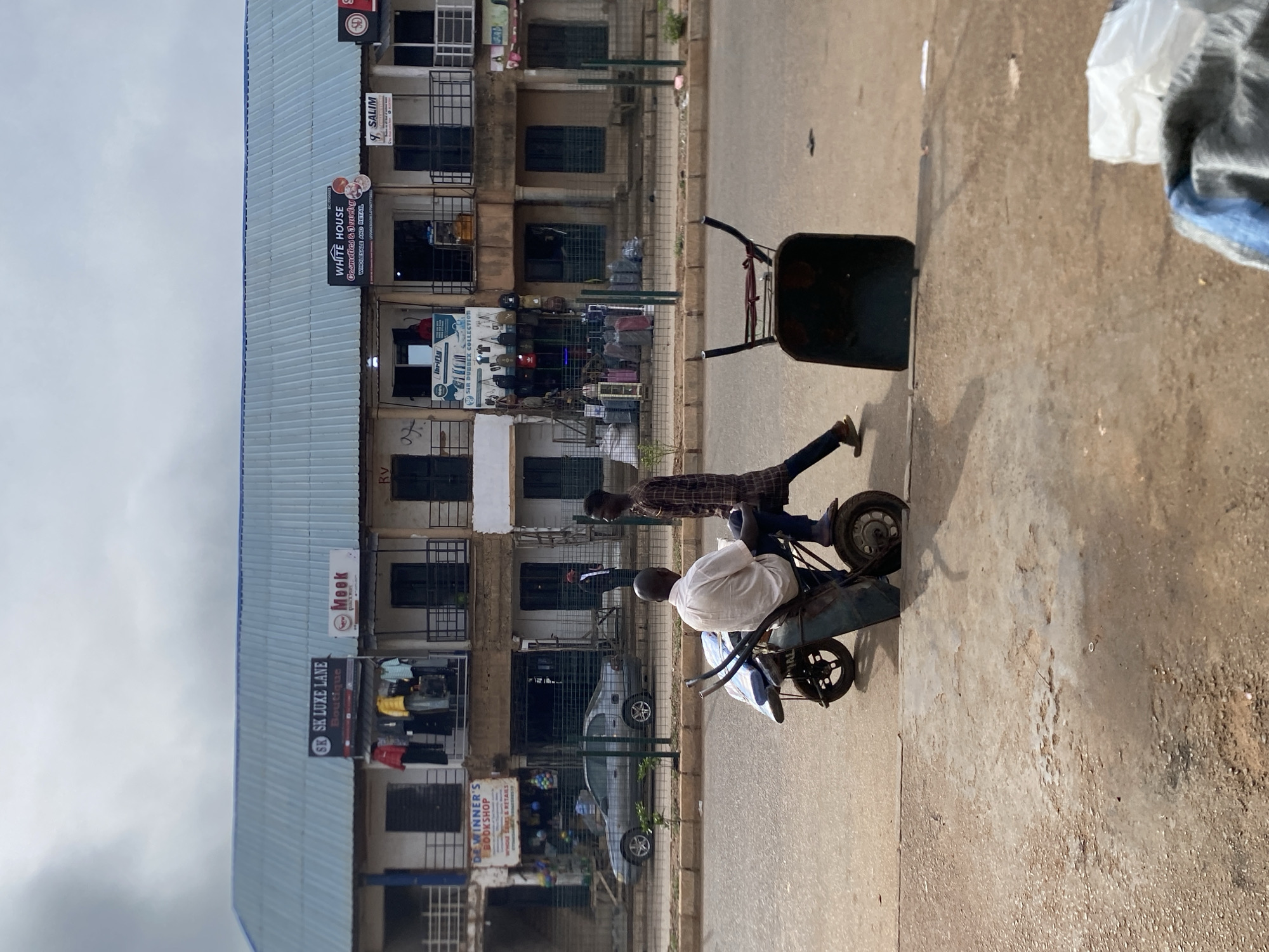The image shows two men moving a two-wheeled cart along a paved road in Bukuru, Nigeria. The man in front is wearing a white shirt and is seated in the cart, while the man behind him is standing and pushing it. The road is lined with multi-story buildings with businesses, including "SK Luxe Lane Boutique", "Mook", "Sir Dubbex", "White House Cosmetics & Jewelry", and "Salim". A car is parked on the road. The sky is overcast.