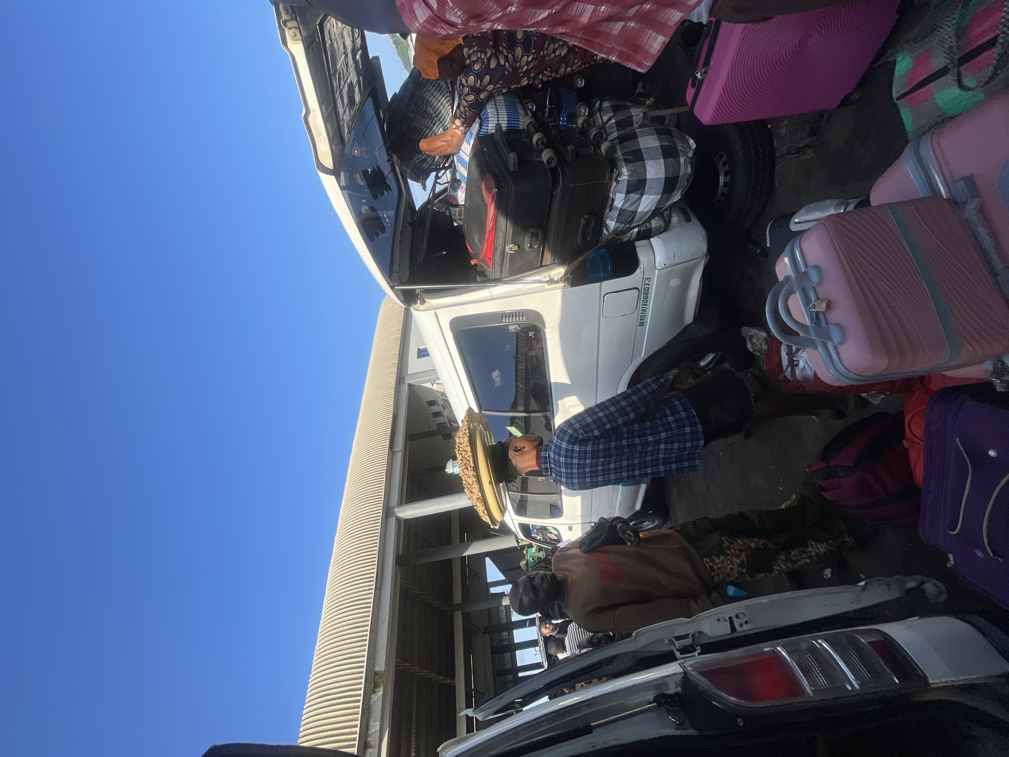 The image depicts a busy scene at what appears to be a transportation hub in Ado-Ekiti, Nigeria. The foreground is filled with luggage, including several pink suitcases, a purple suitcase, and duffel bags. A white van is parked with its rear door open, revealing more luggage packed tightly inside. Two men are standing near the van; one wearing a brown sweatshirt and camouflage pants, the other in a blue and white checkered shirt. A third person, whose face is not fully visible, is also near the van, partially obscured by the luggage. In the background, a covered structure with corrugated metal roofing and concrete supports suggests a market or a bus station. The overall atmosphere is one of transit and commerce, with people preparing to travel or engage in trade. The sky is clear and blue, indicating fair weather.