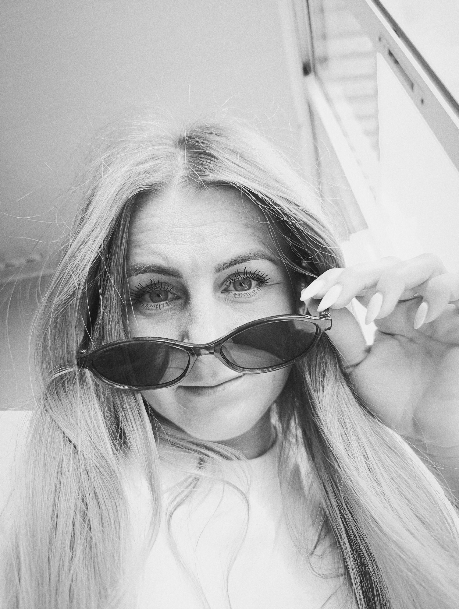 A black and white, close-up portrait shows a woman looking directly into the camera from a low angle. She has long, straight, light-colored hair, which frames her face and flows down her shoulders. Her hair appears slightly windblown or tousled. She is peering over a pair of dark, retro-style sunglasses, which she holds with her left hand (viewer's right). Her eyes are wide and focused on the viewer, with visible eyelashes, and her left eyebrow is slightly raised. A subtle smile plays on her lips. Fine lines are visible around her eyes and on her forehead. Her hand, holding the sunglasses, features long, pointed, light-colored (likely white) manicured nails. The background is blurry and out of focus. On the left side, it appears to be a light-colored wall or surface, while on the right, there are vague hints of a window or architectural structure, suggesting natural light. The overall mood is engaging and playful. There is no visible text in the image.