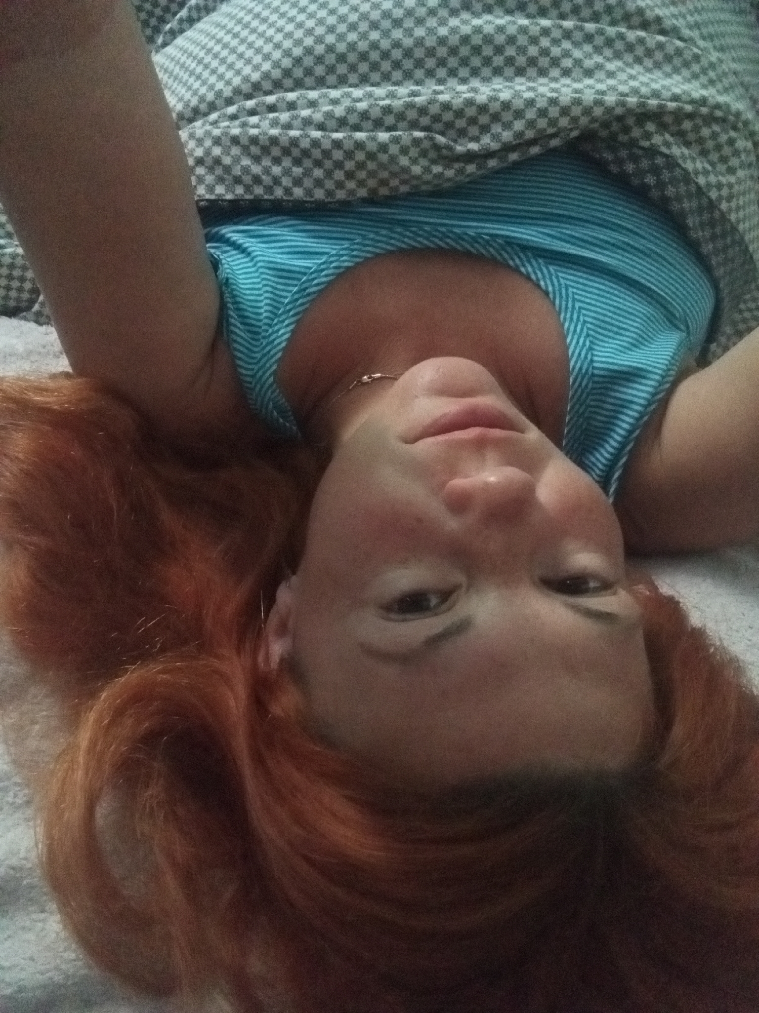 In an indoor setting, a woman with long, auburn hair is lying on a bed, with her head angled toward the viewer. She is wearing a blue and white striped tank top. A grey and white checkered blanket is partially covering her. The scene appears to be a casual, personal moment captured in a home environment, possibly in the morning or evening, given the relaxed posture. There is no indication of the city or location.