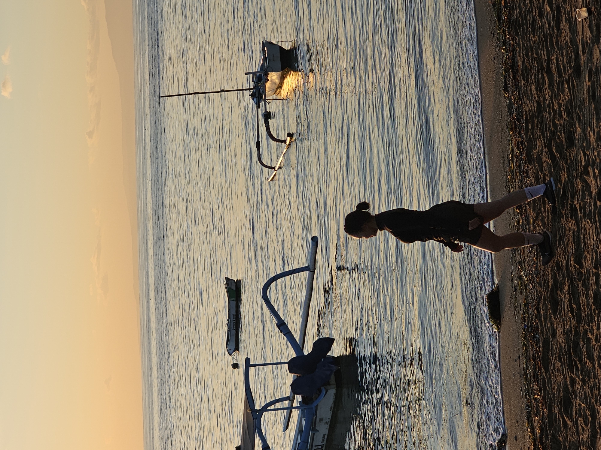 A horizontally oriented, medium shot captures a beach scene, possibly at sunset. The sky transitions from a light orange at the horizon to a pale blue overhead. The sea, mirroring the sky, is marked with gentle waves. A small sailboat is anchored some distance from the shore. In the foreground, a woman in dark clothing stands on the sandy beach. Her hair is tied in a bun. A blue metal structure stands in the water nearby. The sandy beach transitions into darker ground. The location might be in Denpasar, Indonesia.