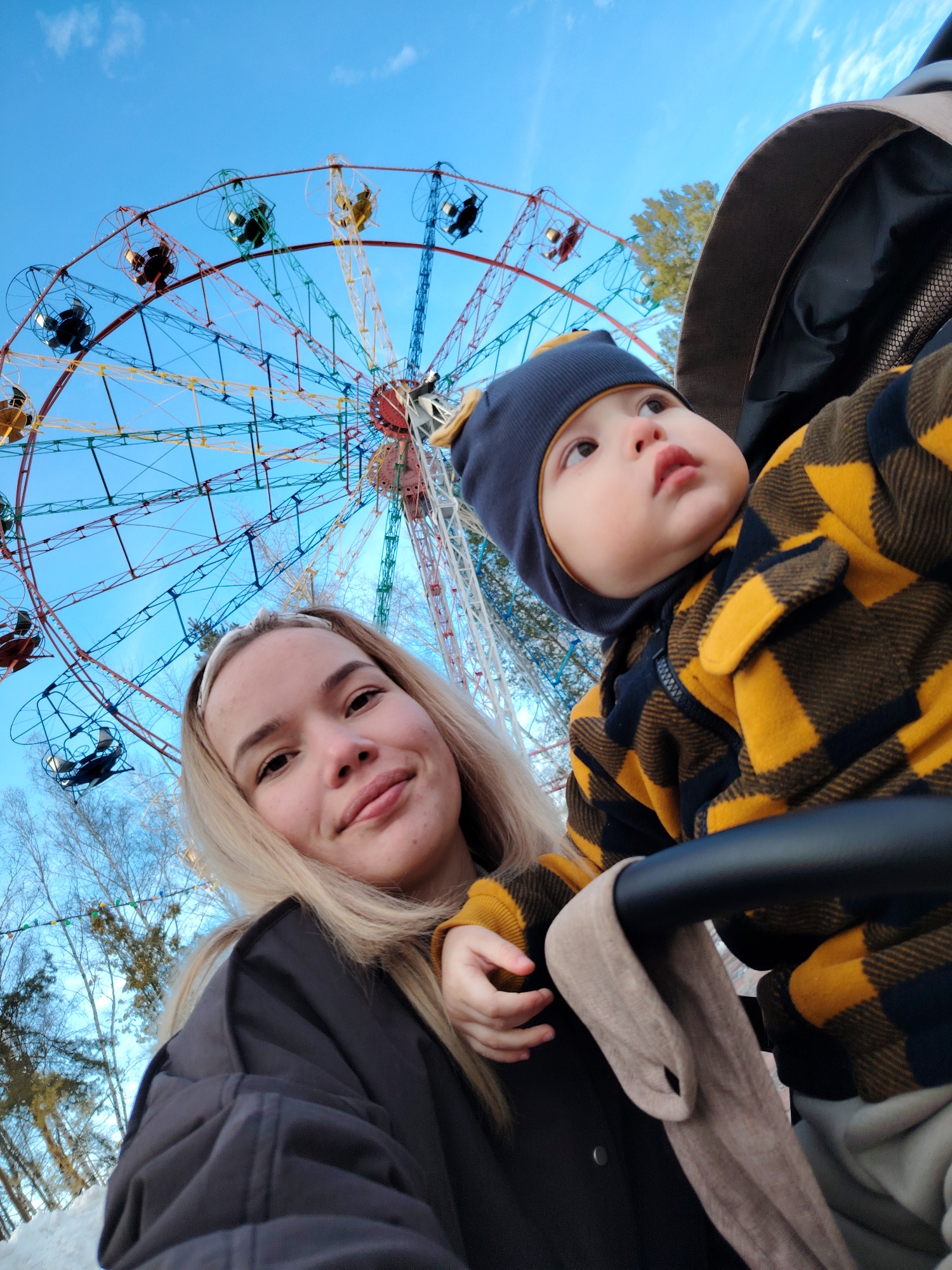 An outdoor scene in an amusement park in Chaikovsky, Russia, depicts a young woman and an infant. The environment suggests winter or early spring conditions, with snow visible on the ground and bare deciduous trees in the background, set against a clear blue sky with scattered clouds. A large, multicolored Ferris wheel, featuring red, blue, yellow, and green structural elements and numerous open-air gondolas, dominates the upper background.
The woman, with long blonde hair and a white headband, occupies the lower-left to central foreground. She wears a dark jacket and looks directly at the camera with a neutral expression. To the right of the woman and slightly elevated, an infant is seated in a dark-framed stroller or carrier, identifiable by its dark handle and tan fabric. The infant wears a dark blue beanie with yellow accents and a yellow and black plaid zip-up jacket. The infant's gaze is directed upwards and to the right, away from the camera. A portion of the woman's left hand is visible near the infant's arm.