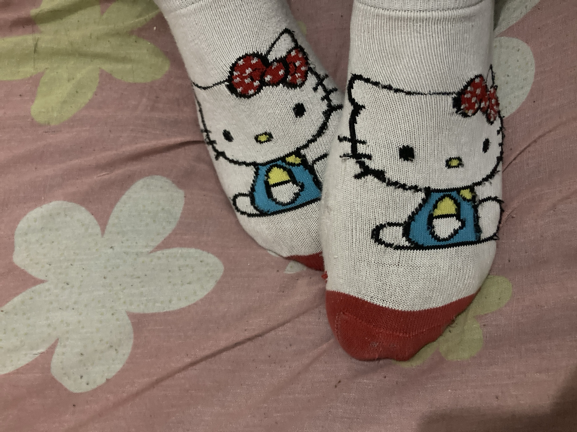 The image captures a close-up, high-angle view of two feet adorned with white socks, resting on a patterned fabric surface. The socks prominently feature an embroidered or printed design of Hello Kitty. Each Hello Kitty character is depicted wearing a blue dress or overall with yellow accents, and a distinctive red bow with white polka dots on her left ear (from the viewer's perspective). The heels and toes of the socks are a contrasting red color. Slight wear, such as minor fraying, is visible along the black outlines of the Hello Kitty design, particularly noticeable on the sock to the right.
The background is a pink textile, likely a bedsheet or blanket, decorated with large, stylized floral patterns in light green and white. The fabric itself shows signs of use, including some pilling and small dark specks that could be lint or dust. The setting appears to be indoors. The lighting is soft and diffused, suggesting either artificial indoor lighting or indirect natural light. There is no specific activity taking place, only the feet resting. No visible text is present in the image.