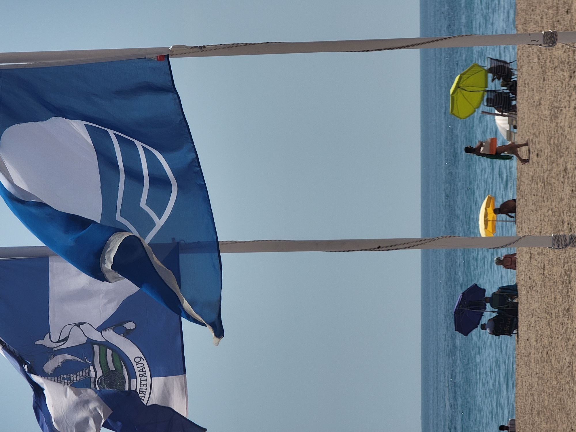 The image captures a sunny day on the beach in Quarteira, Portugal. Two large blue and white flags are prominently displayed in the foreground, fluttering in the breeze. The flag on the left features a crest with a ship and the word "QUARTEIRA" written in reverse. The flag on the right has a stylized white design. In the background, the blue sea stretches to the horizon under a clear sky. The sandy beach is populated by a few beachgoers. Two people are seated under a dark blue umbrella, and another person is walking along the shore with a bag. Several beach umbrellas, including a bright yellow one and a lime green one, are dotted across the sand. The overall scene depicts a relaxed beach environment in Quarteira.
