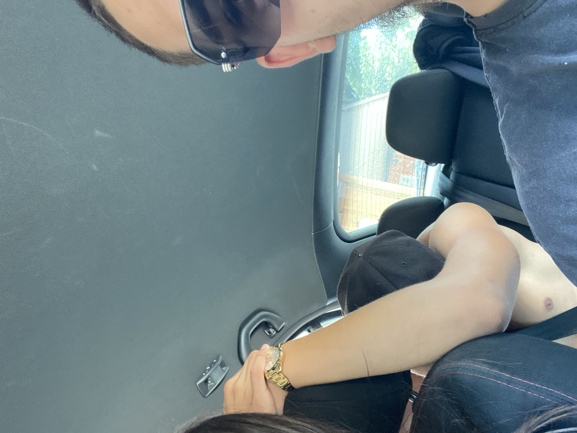 The image shows the interior of a car, likely a four-door sedan, with a focus on passengers and the car's ceiling. The roof lining is dark gray and textured, with a small overhead light fixture visible towards the top left. The car's interior trim is also dark. On the left side of the frame, a person's arm is visible, wearing a gold watch. This arm is bent, supporting what appears to be another person's head, who is wearing a black baseball cap. To the right, a person wearing sunglasses is visible, with their right ear and side of their head in frame. The person is wearing a dark t-shirt. The background shows the rear seats of the car, also in dark upholstery, and a window revealing greenery and buildings outside, suggesting an outdoor setting, possibly in a residential area. The specific location of Hirevo, Bulgaria, cannot be determined from the visual information in the image.