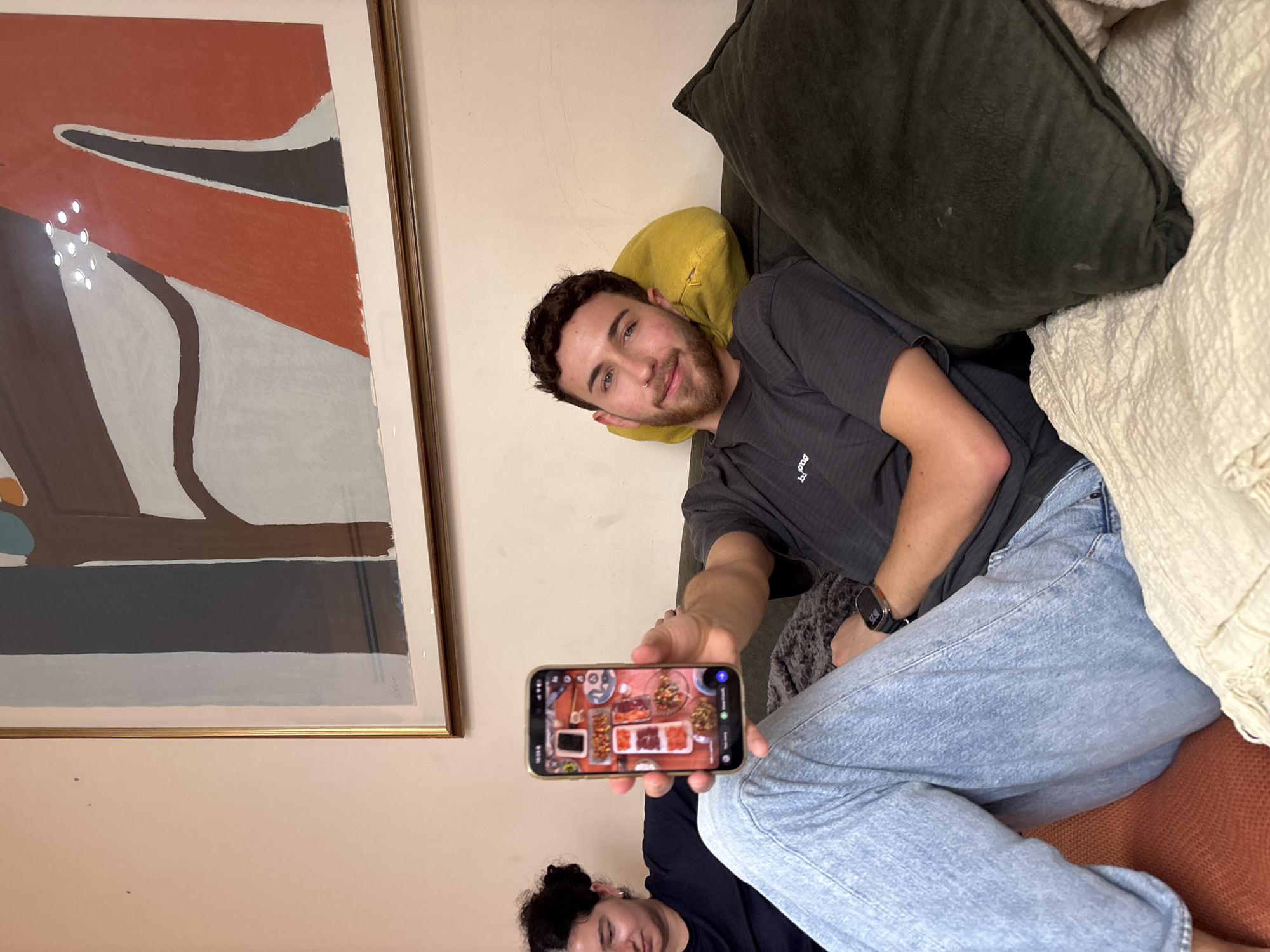 A man is lying on a couch in an indoor setting, possibly an apartment in Tel-Aviv, Israel. He is holding a smartphone displaying an image of a food platter. Another person's head is visible in the lower portion of the image. The couch has several pillows. There is a large framed piece of art hanging on the wall. The man is wearing a dark t-shirt and light blue jeans, and has a watch on his left wrist.